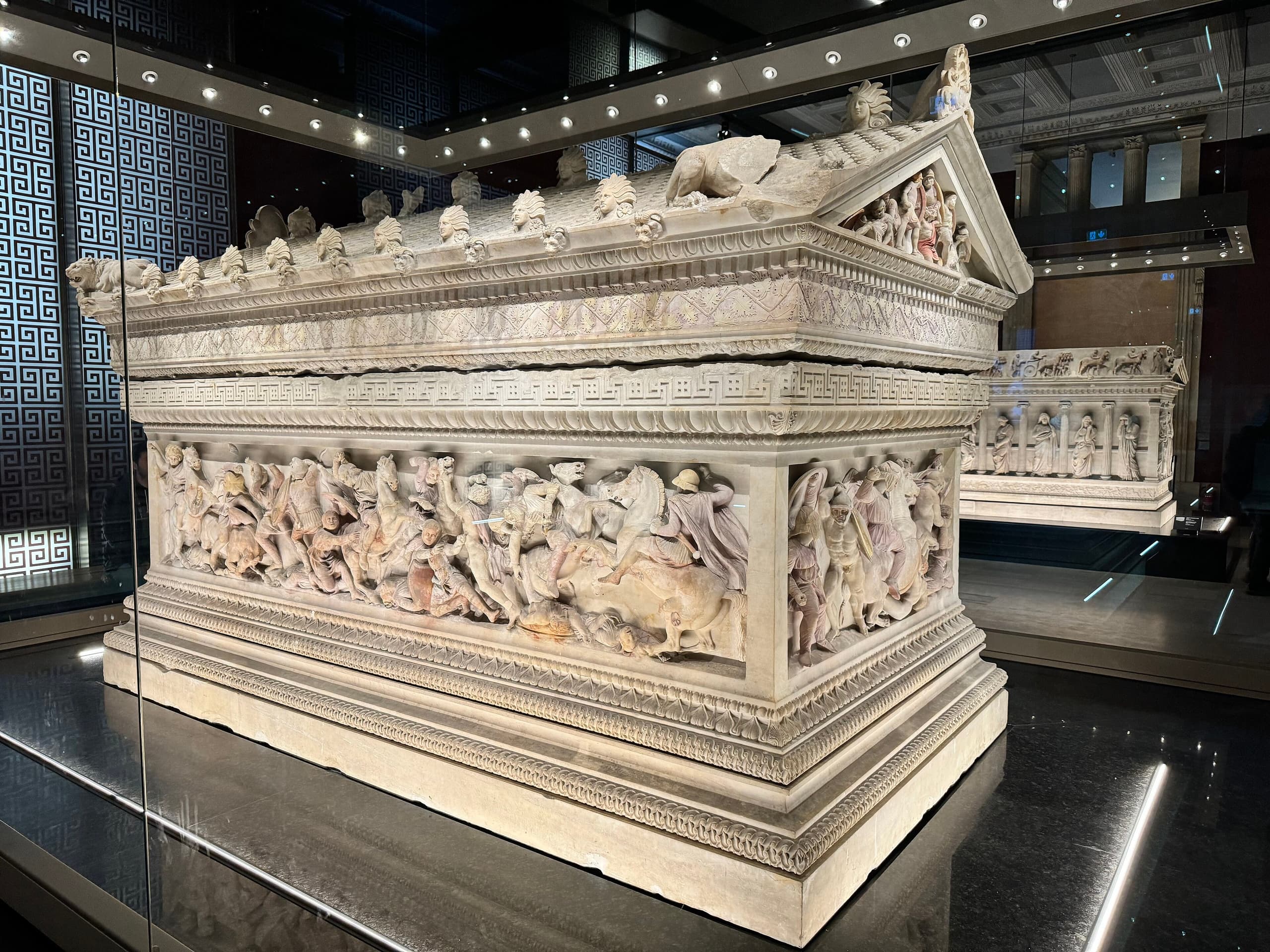 A large ancient marble sarcophagus densely carved in high relief with crowded figures of warriors, horses and attendant scenes along its long side, the stone still showing faint traces of old colour in places.