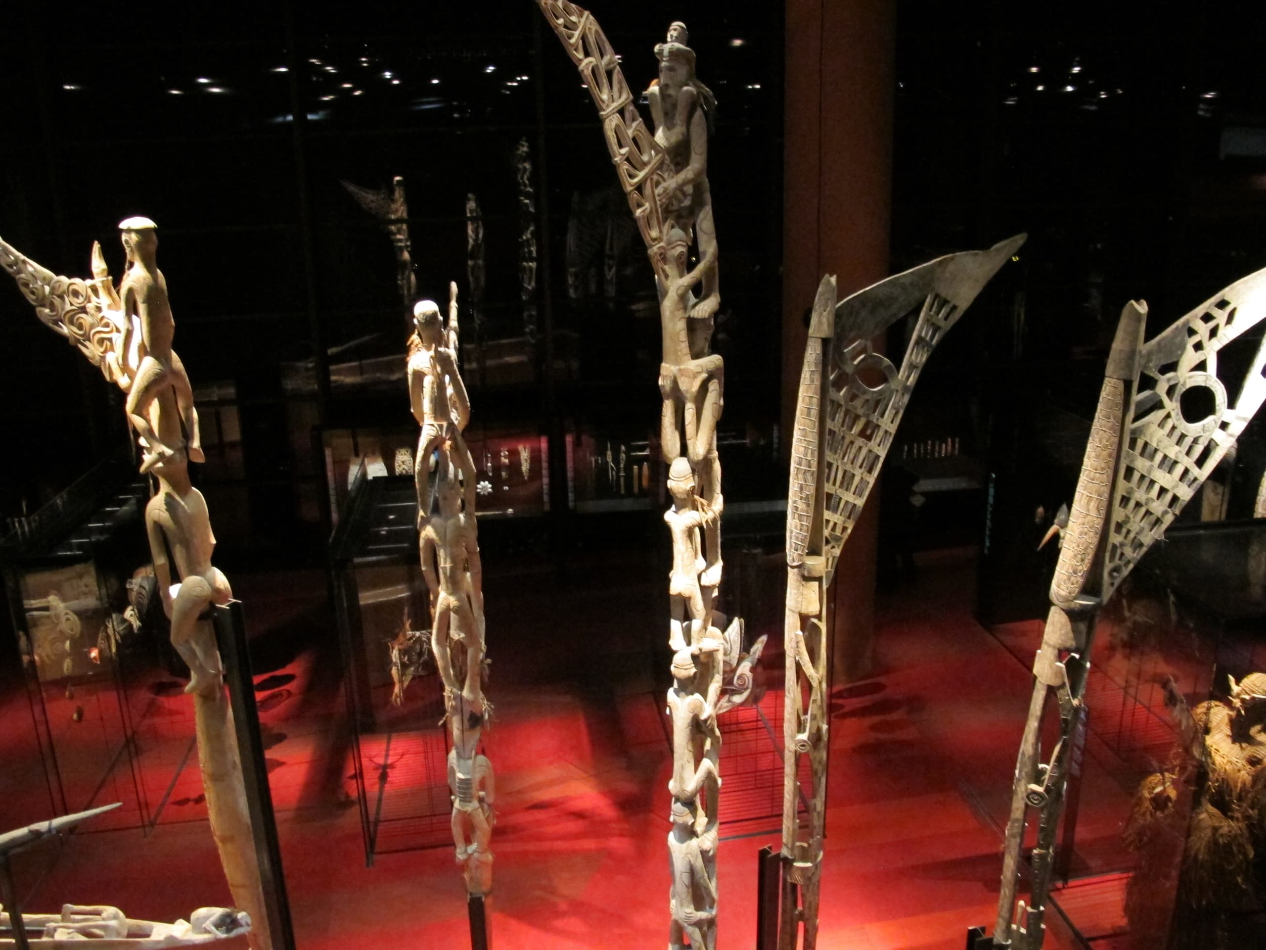 A tall carved wooden pole with stacked human figures along its length and an elaborate openwork projection at the top, painted with white and red pigment.