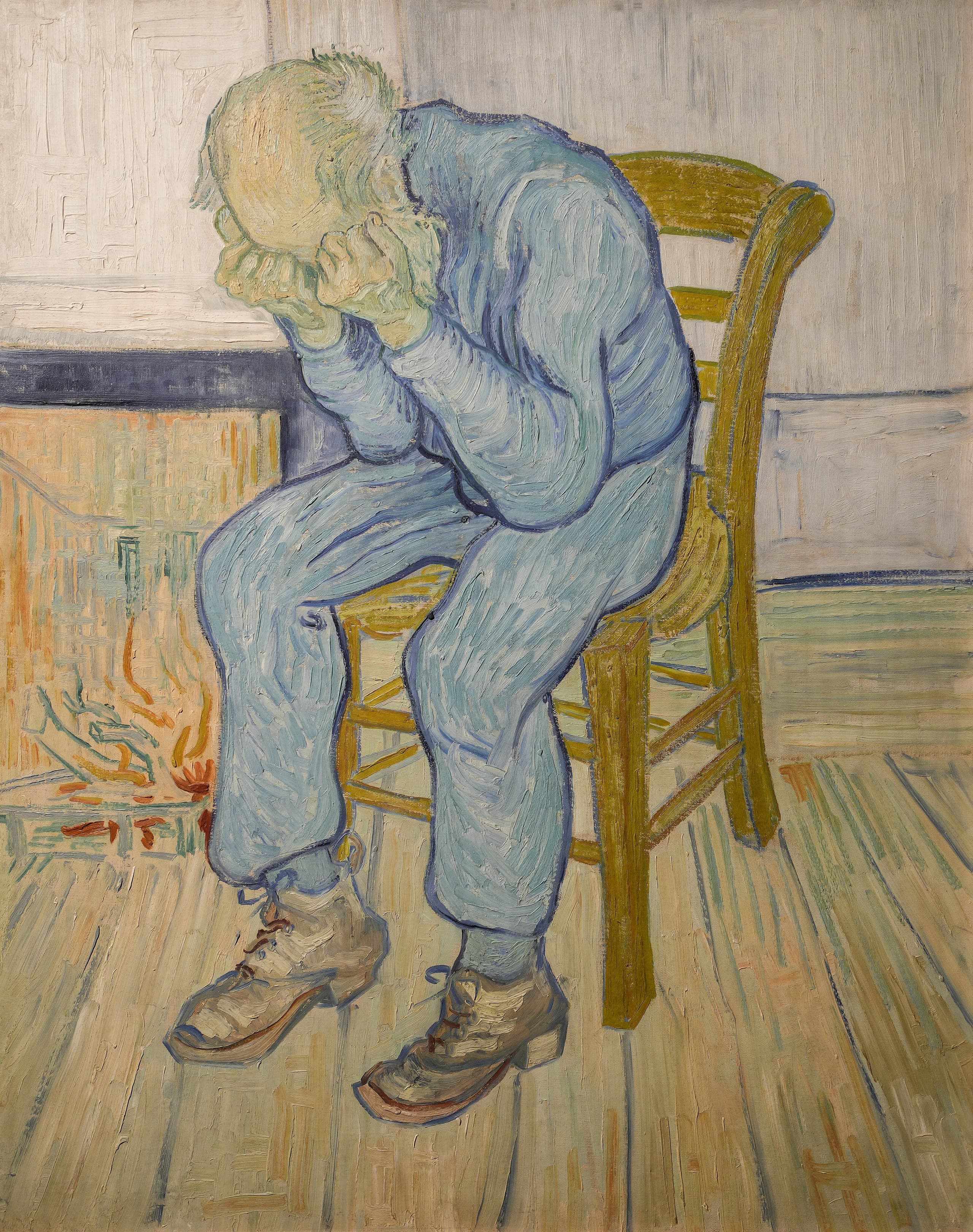 An elderly man in a blue jacket and trousers sits hunched on a wooden chair, elbows on knees, both clenched fists pressed against his eyes so the face is hidden. The room around him is plain and nearly empty.