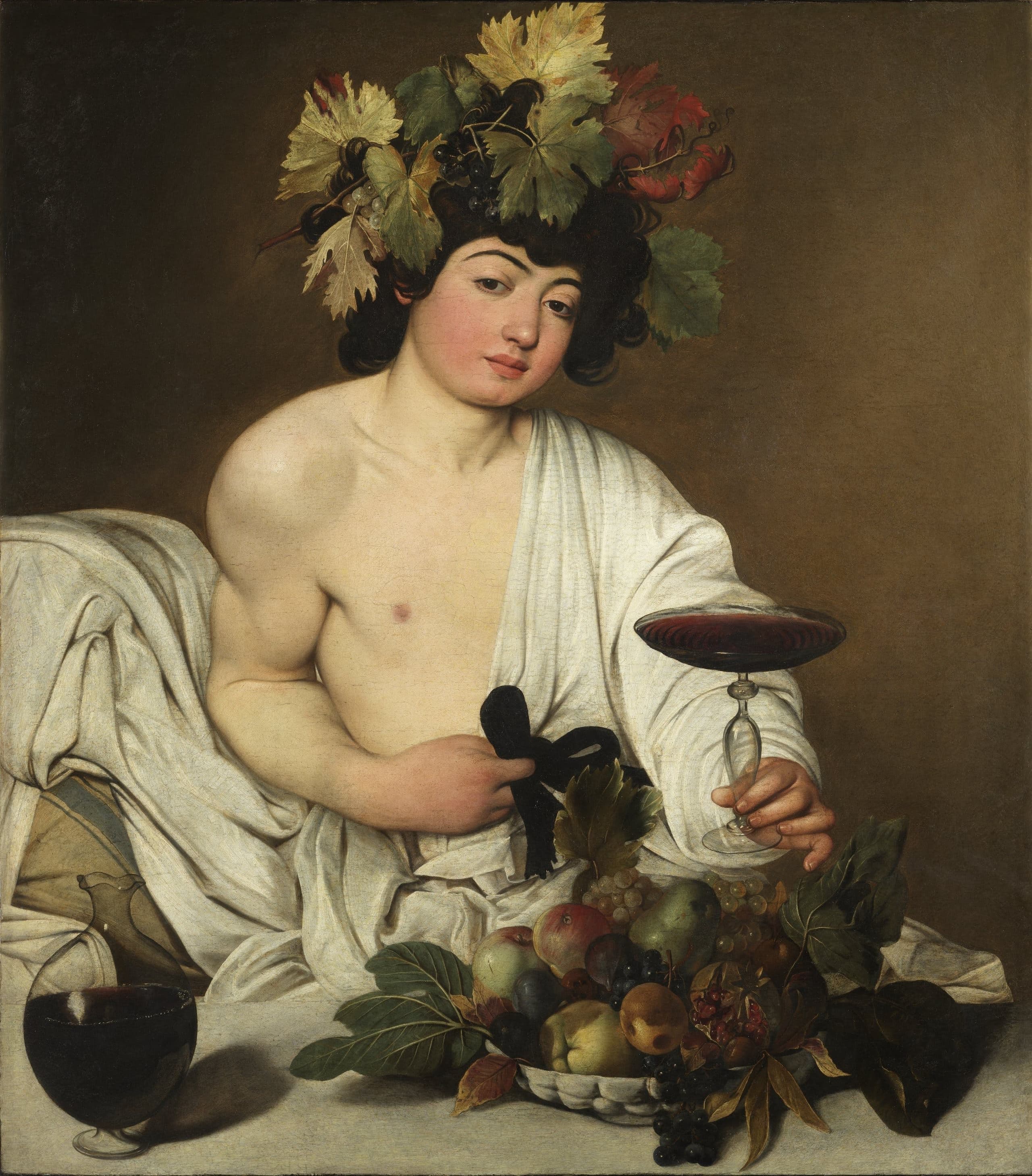 A pale youth with vine leaves and grapes in his dark hair reclines behind a stone table, white robe slipped from one shoulder, holding out a shallow goblet of red wine beside a bowl of fruit and a glass carafe.