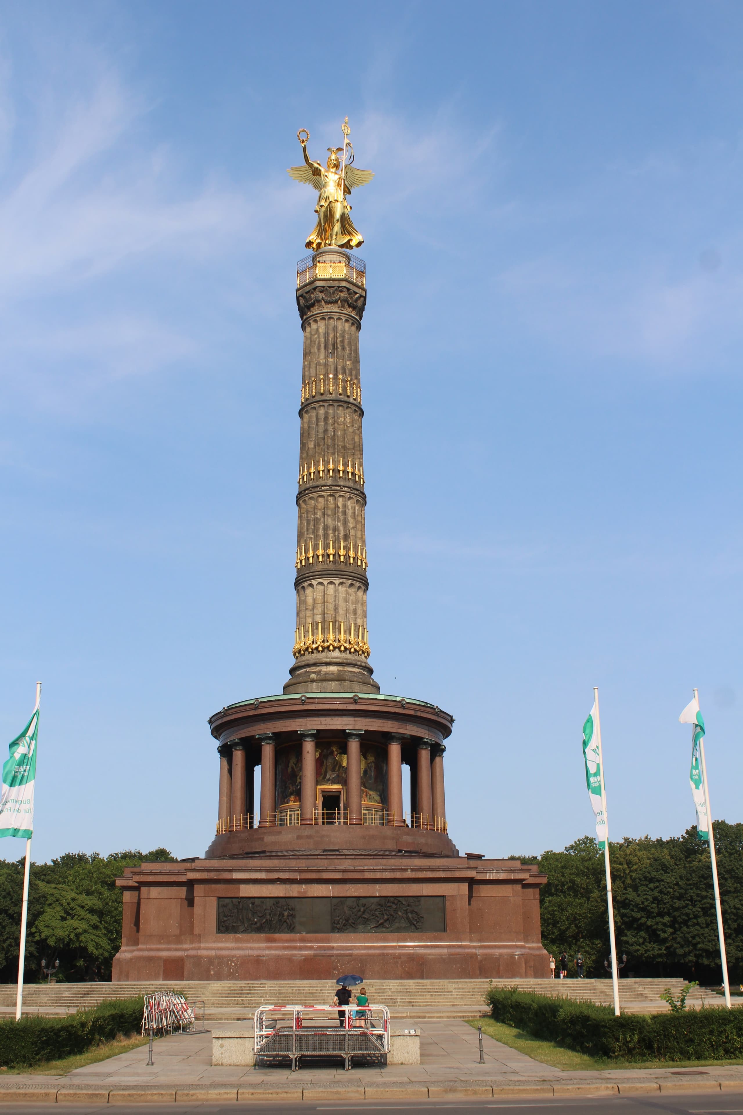 A tall freestanding column in open parkland, broad at the base and rising through stacked stone drums to a gilded bronze figure at the summit.