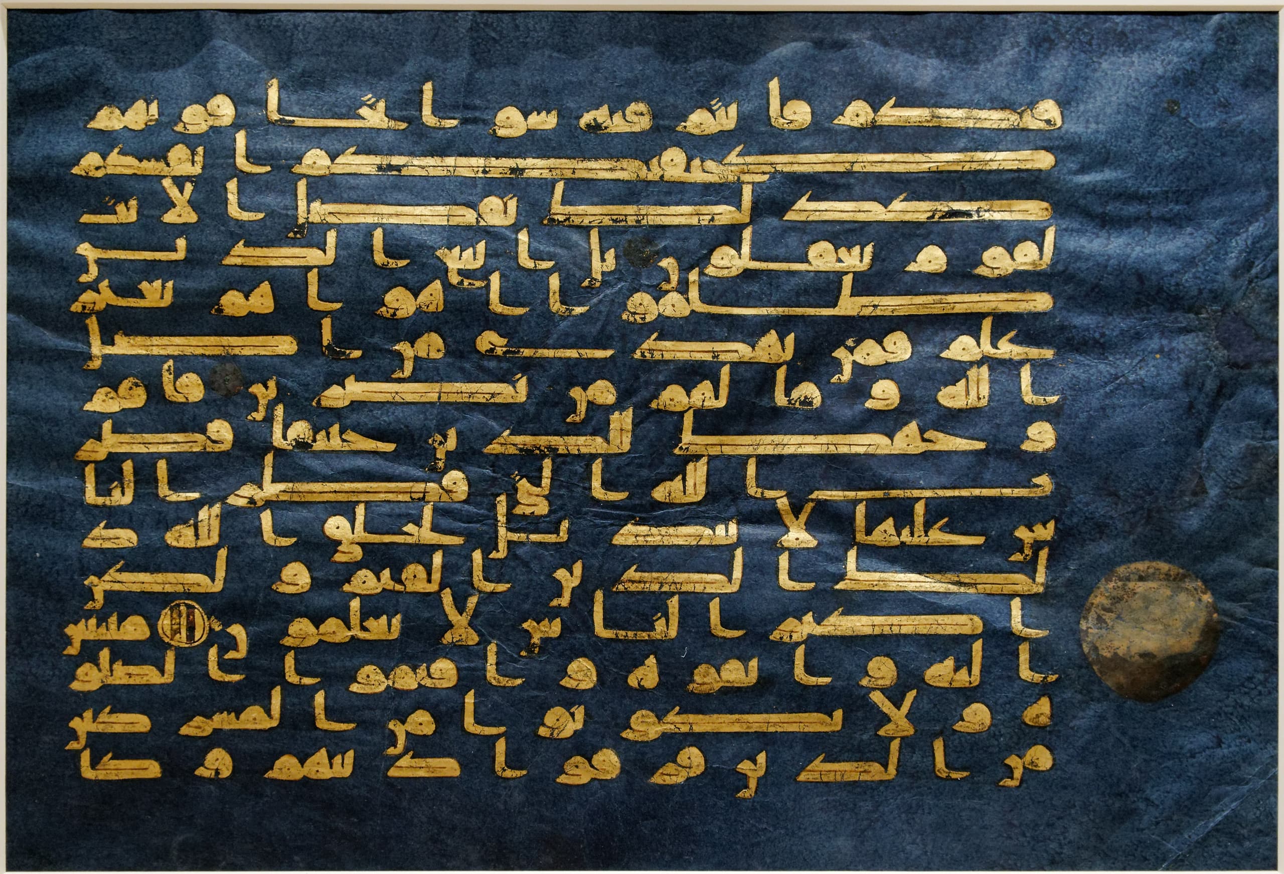 A page of Arabic text in gold on deep blue parchment. The letters are angular and geometric, arranged in horizontal lines with small silver markers between verses.