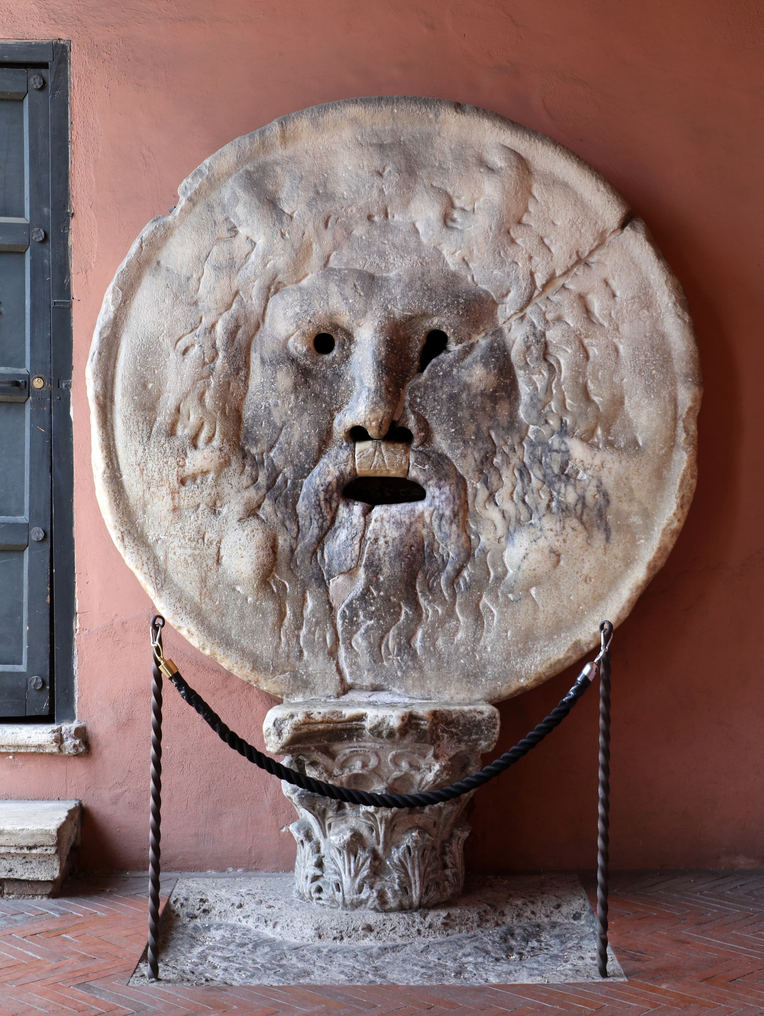 A large circular marble relief of a bearded male face with hollow eyes, open nostrils and a wide open mouth, set against a wall.