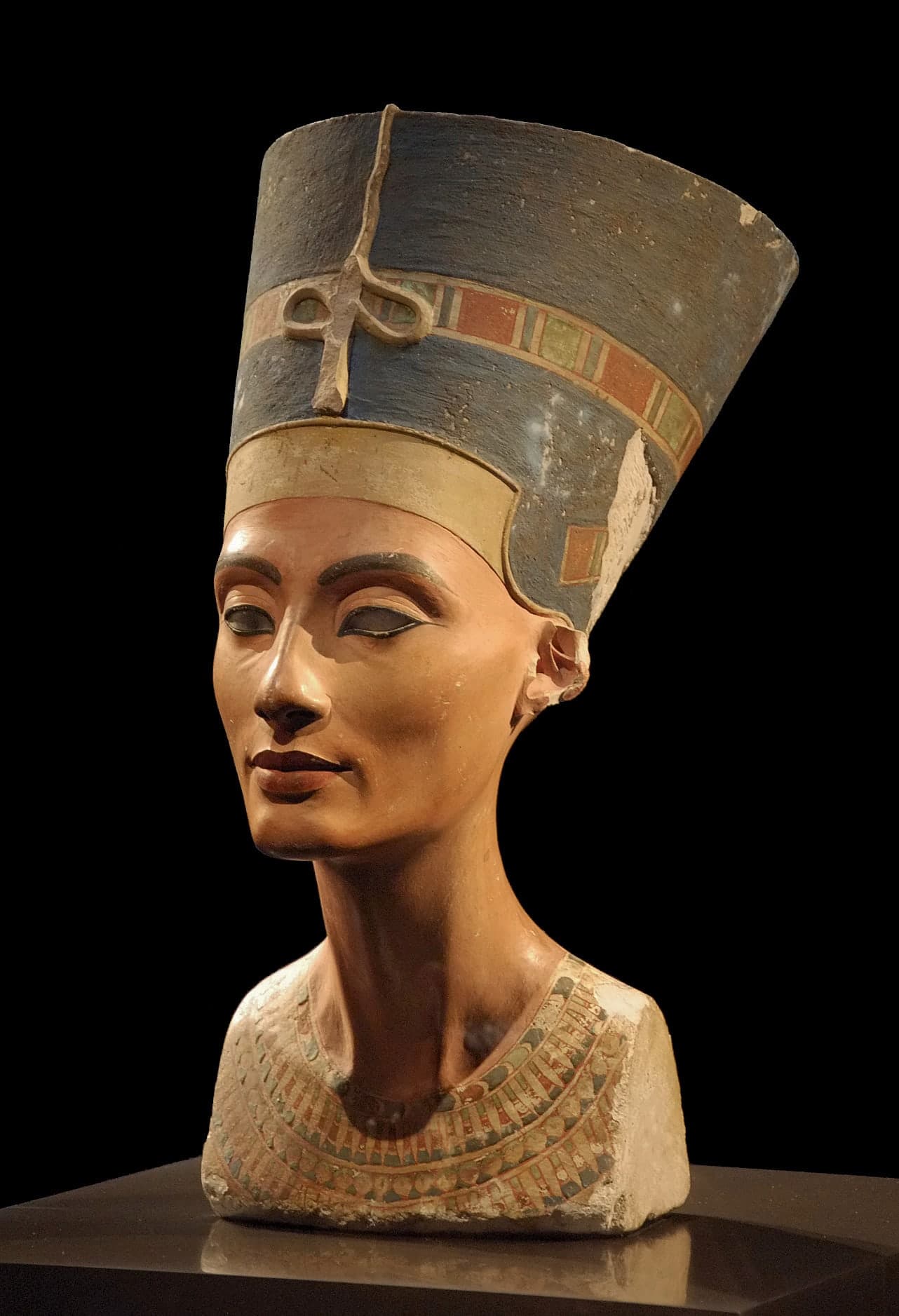 A limestone bust of a woman wearing a tall flat-topped blue crown, with a smooth face, long neck, and one inlaid eye. The left eye socket is empty.