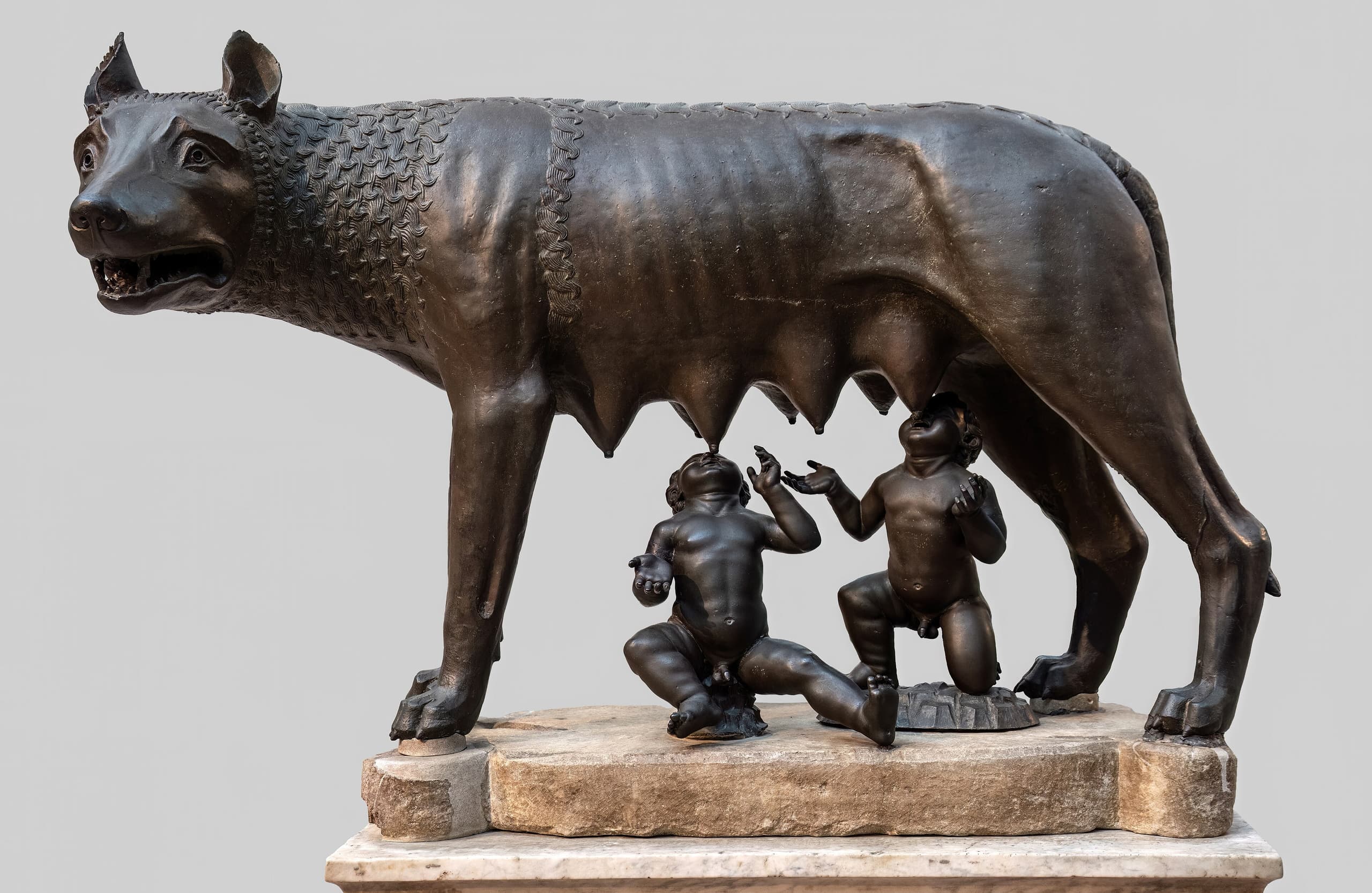 A bronze she-wolf stands in profile, legs planted and mouth open, while two small infant boys sit beneath her belly reaching up to suckle.
