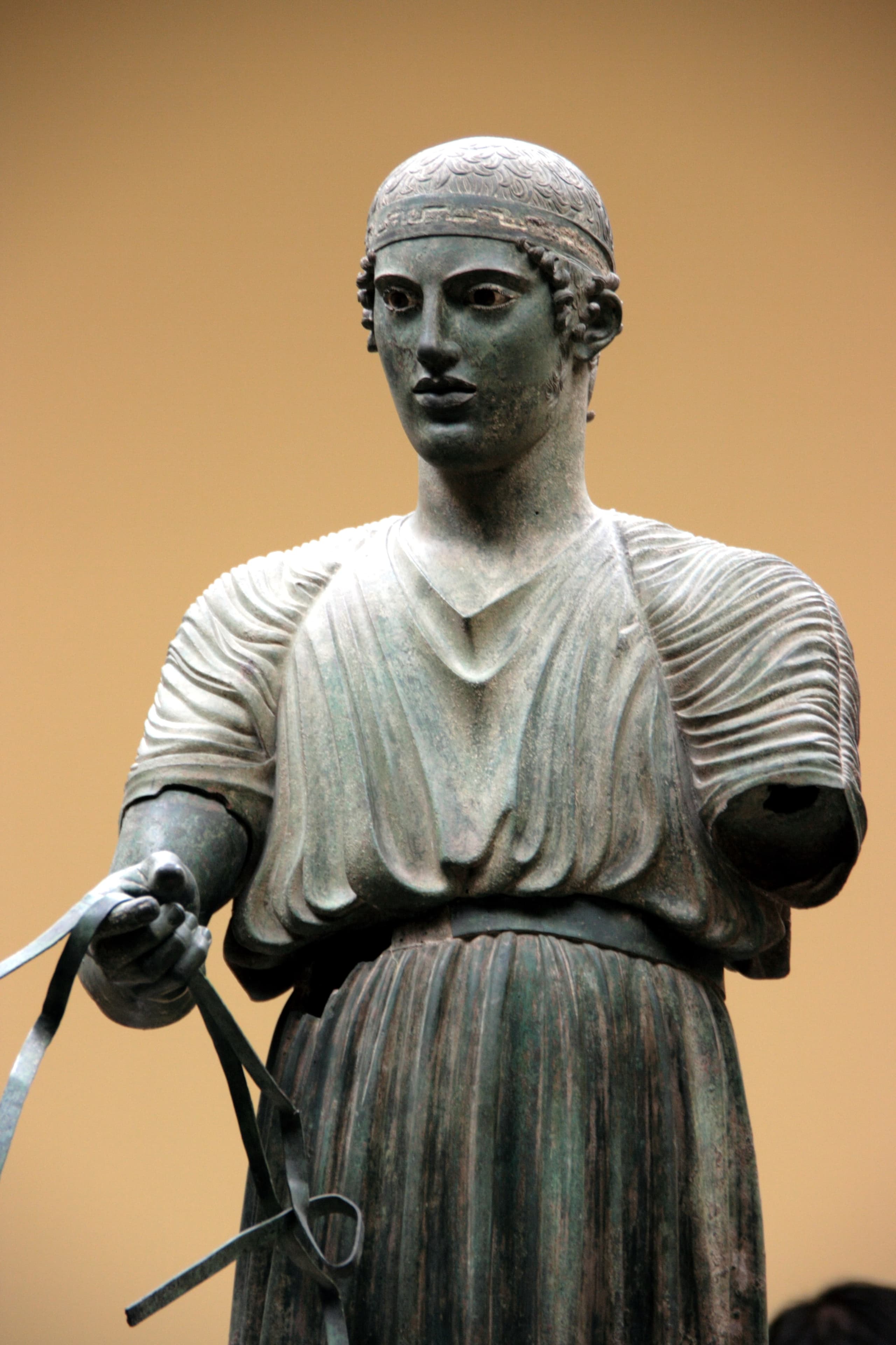 A standing bronze figure of a young man in a long pleated tunic and headband, right arm extended, head turned slightly left, with inlaid eyes and detailed facial features.