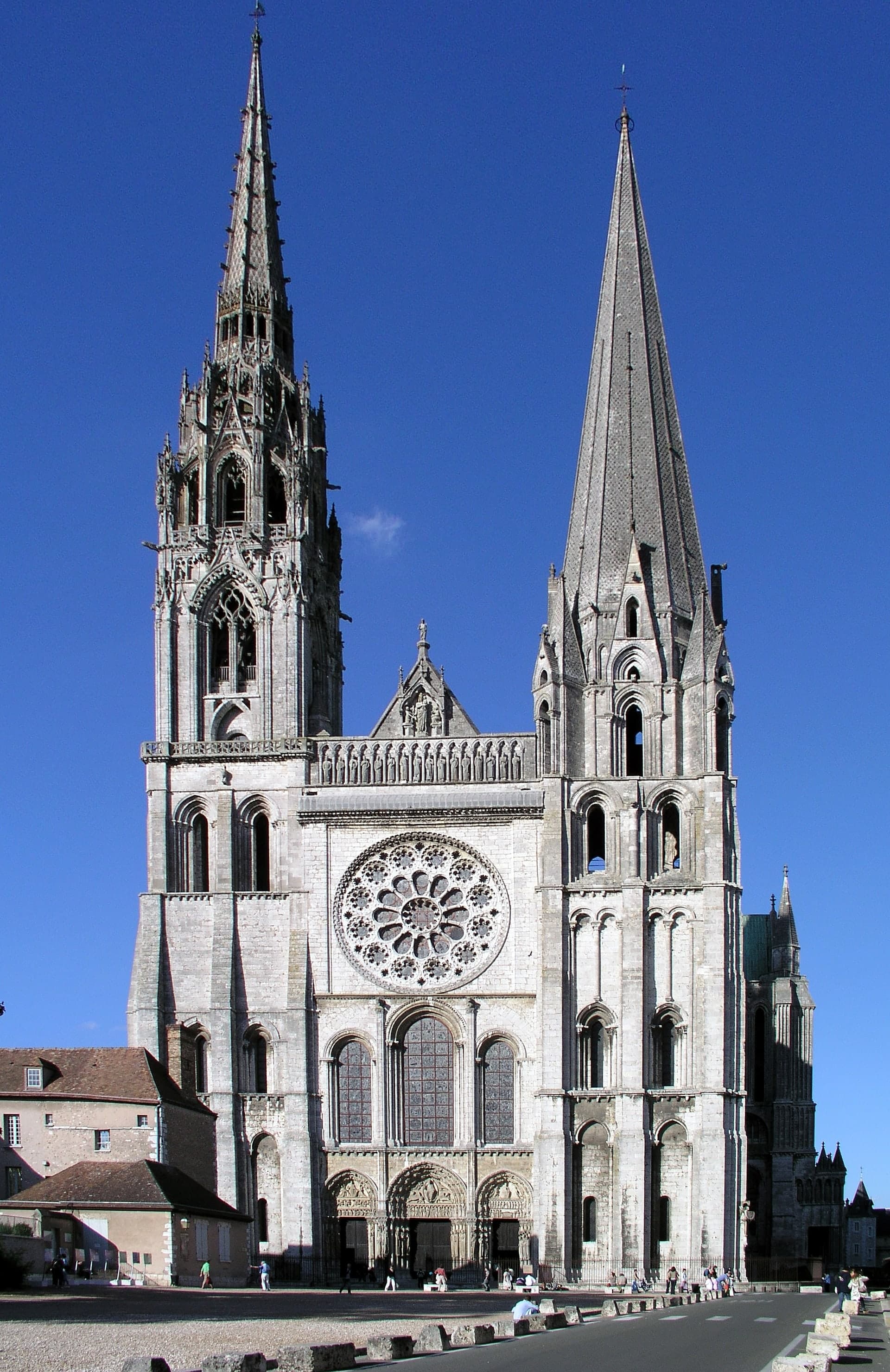 A tall Gothic cathedral with two unmatched spires, one plain and one ornate, and a façade filled with large rose windows and pointed arches.