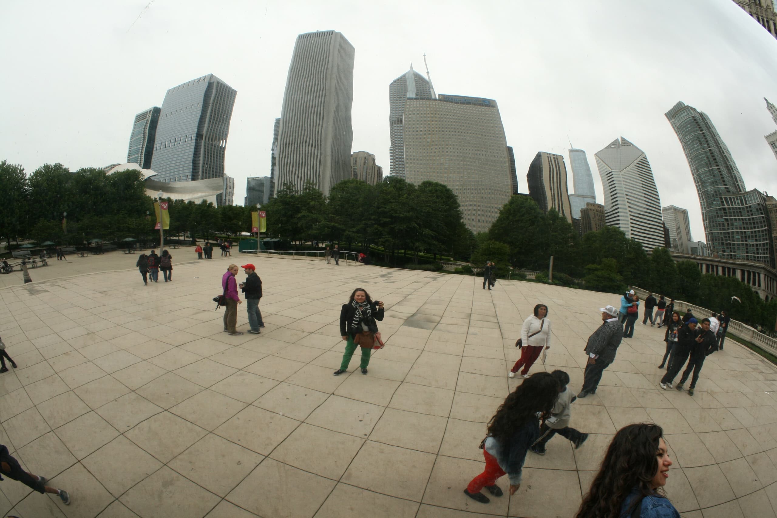 A large, smooth, bean-shaped steel sculpture in a plaza, its mirror surface reflecting buildings and sky, with an open arch beneath and people walking around it.