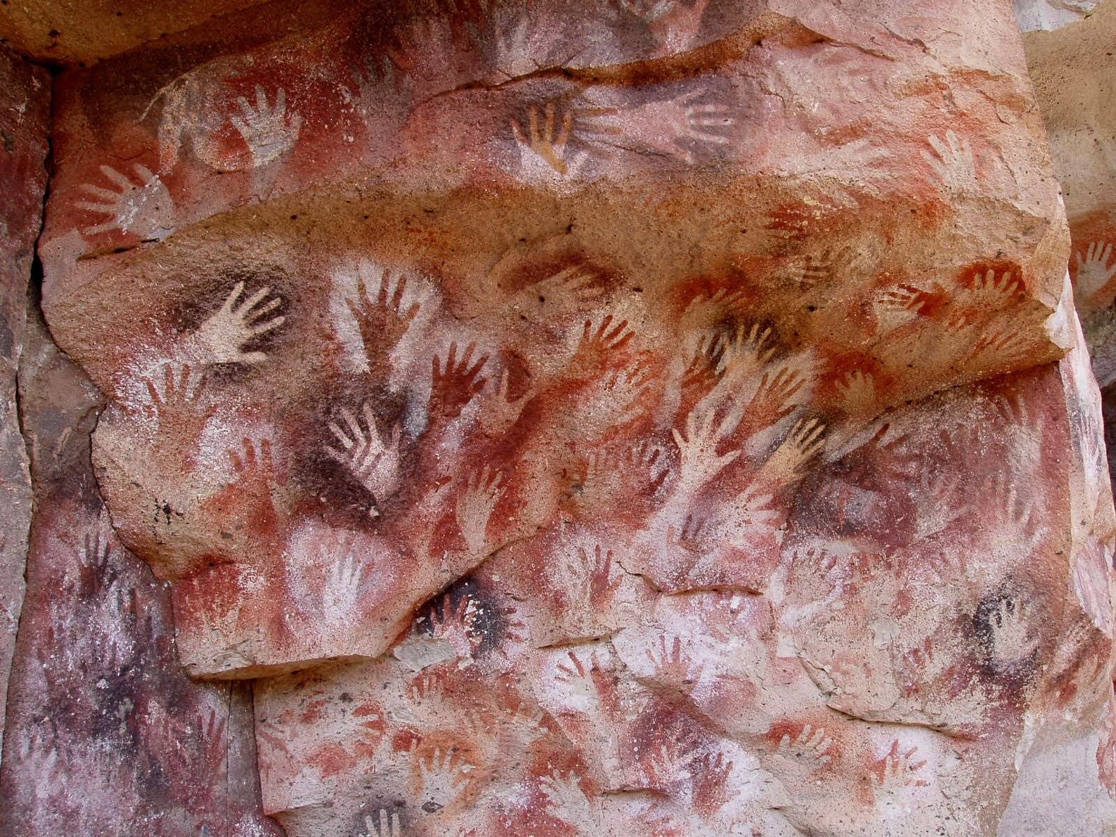 A rock wall covered in overlapping hand stencils in red, orange, and purple pigment, with the hands themselves appearing as unpainted negative space against the coloured background.
