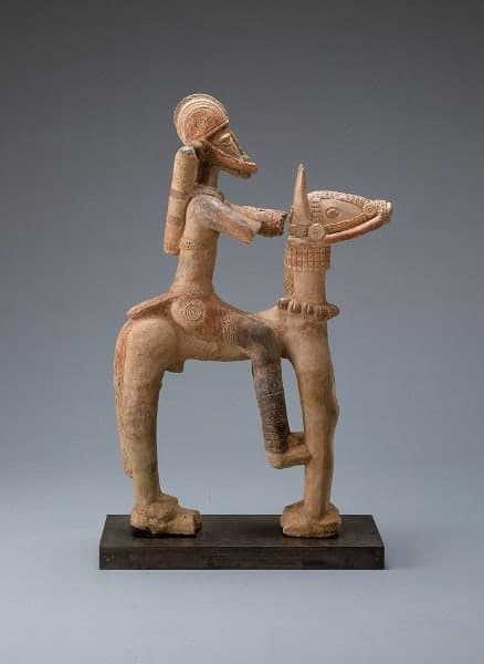 A kneeling terracotta figure with arms wrapped around the torso, raised scarification lines on the face, and serpents coiled along the back and shoulders.