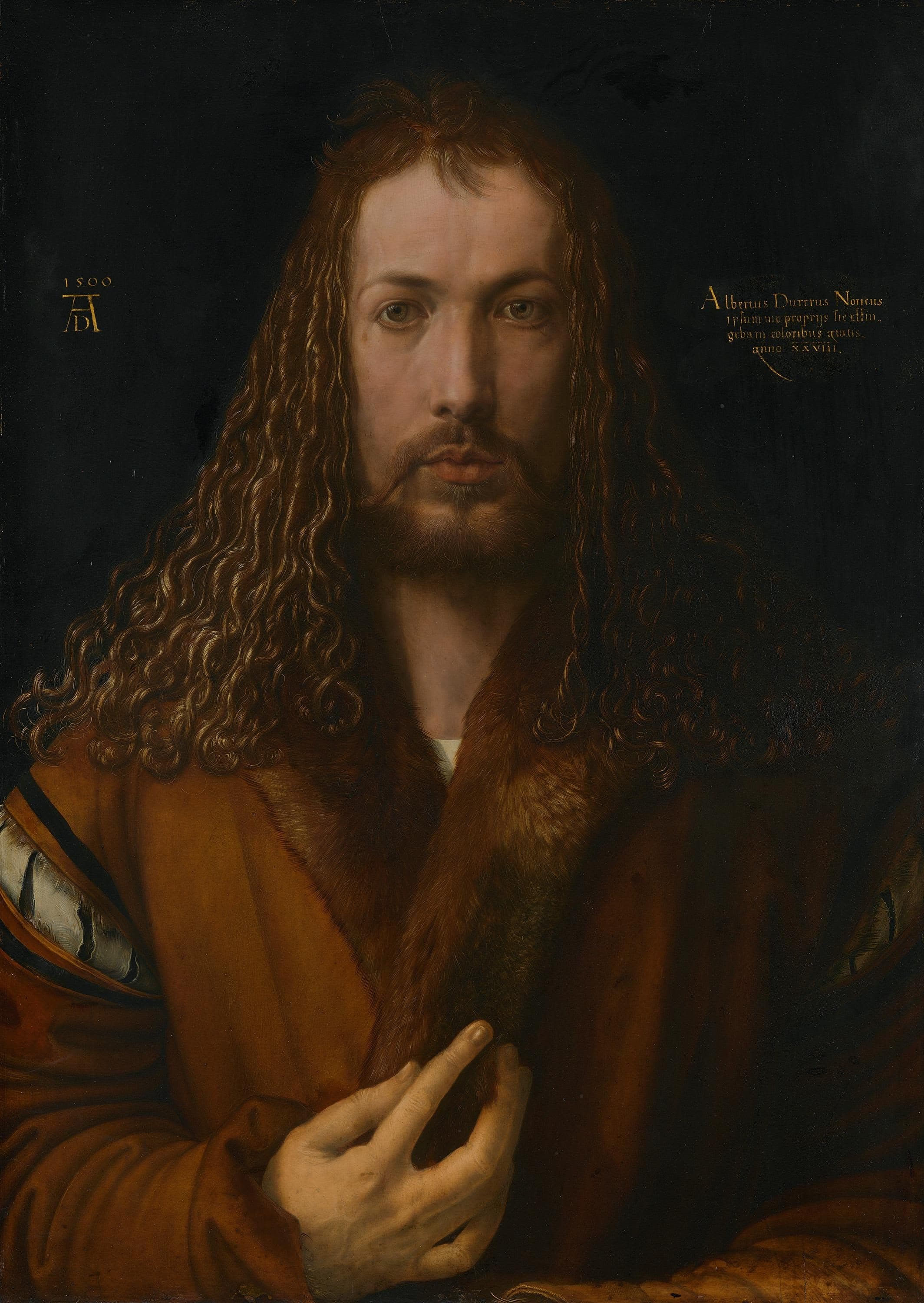 A man with long dark curly hair and a dark fur-trimmed robe, facing forward, right hand raised near the centre of his chest, against a plain dark background.