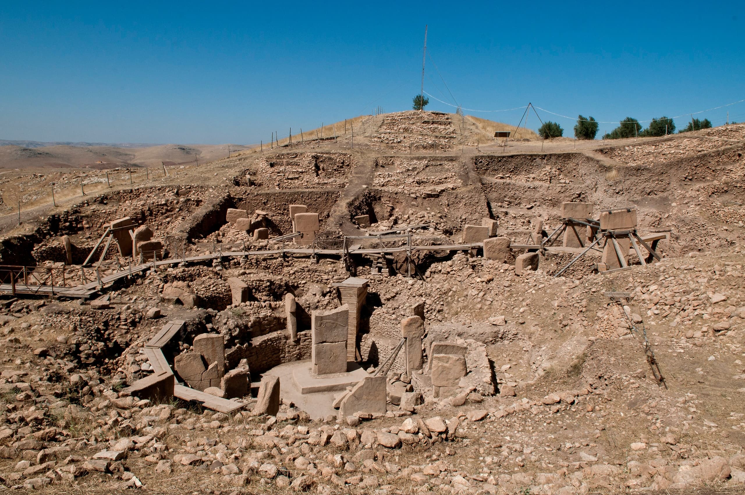 Excavated stone pillars standing in a circular arrangement on a hillside. The pillars are T-shaped, carved from pale limestone, with animal figures visible in relief on some surfaces.