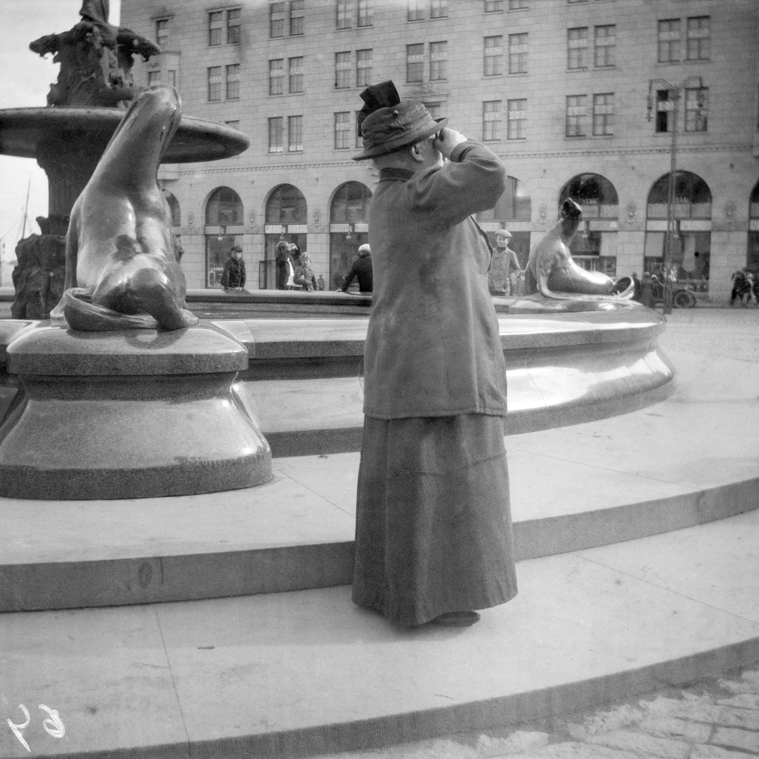 A tall dark bronze fountain sculpture in an open square, the standing figure raised high above a lower basin level where the fountain elements sit.