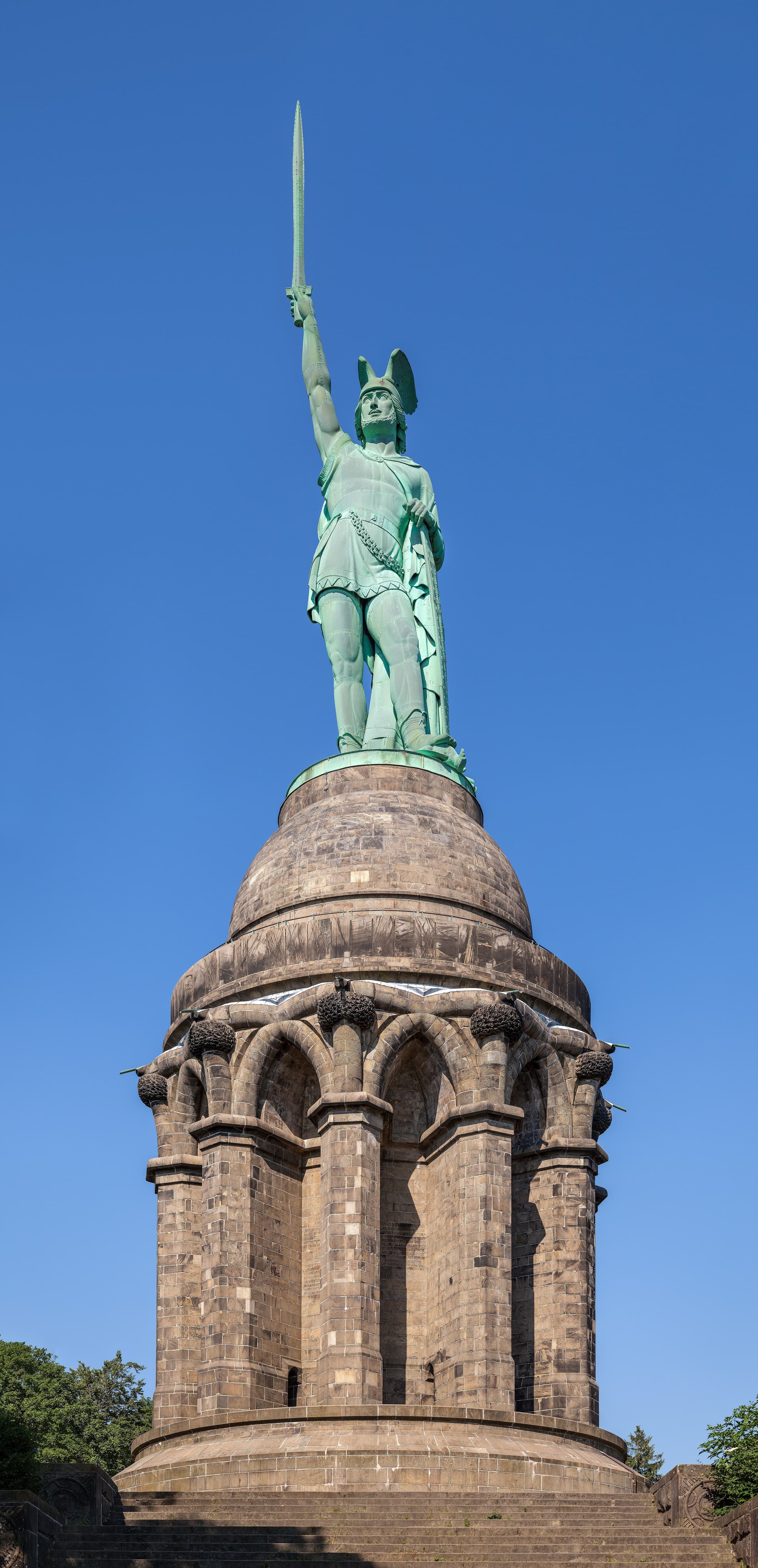 A colossal iron statue of a warrior standing on a tall cylindrical stone pedestal, right arm raised holding a sword pointed skyward, surrounded by dense forest.
