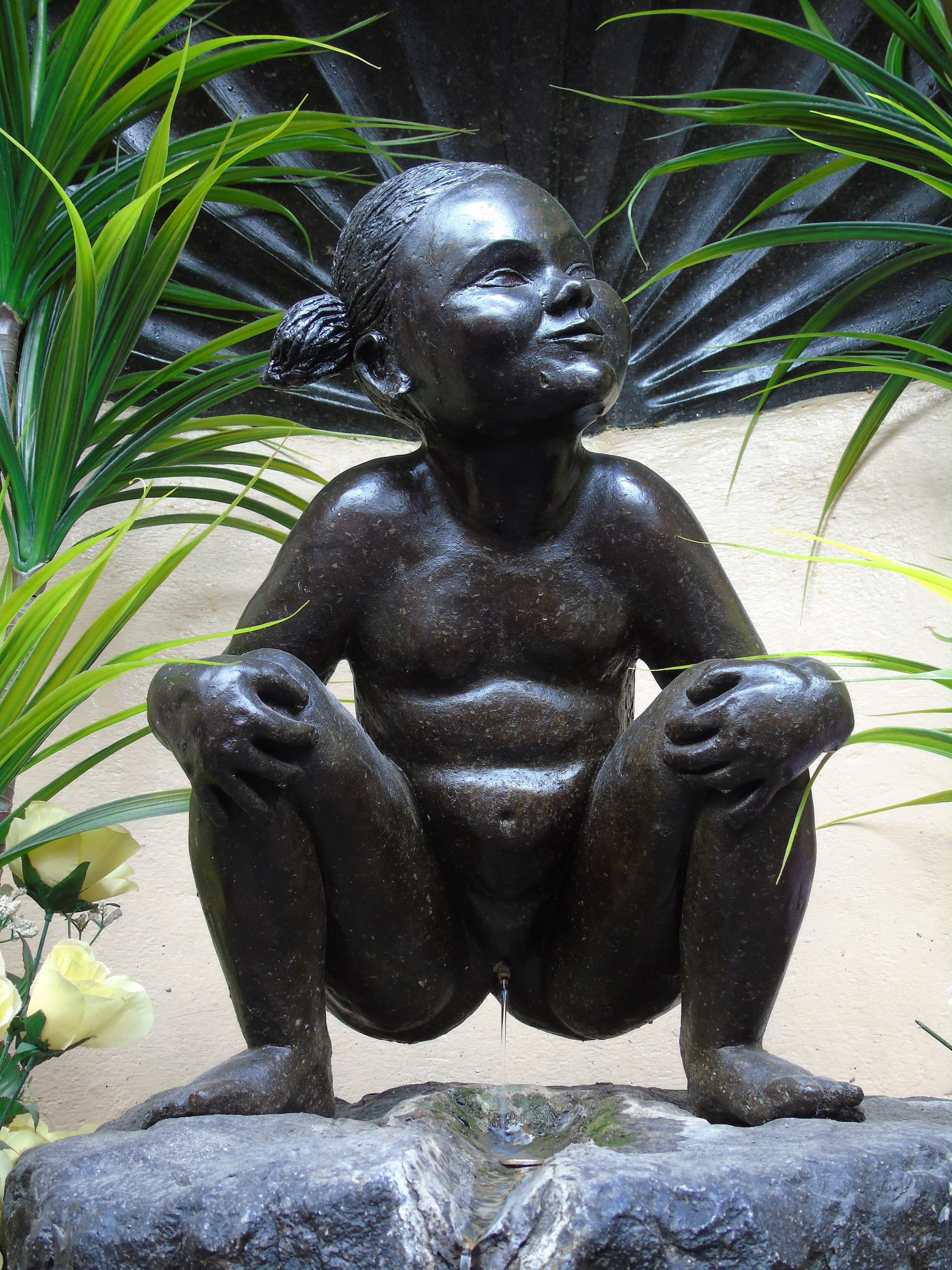 A small bronze figure of a naked girl with short pigtails, squatting over a fountain base as if urinating, seen at close range and partly framed by protective iron bars.