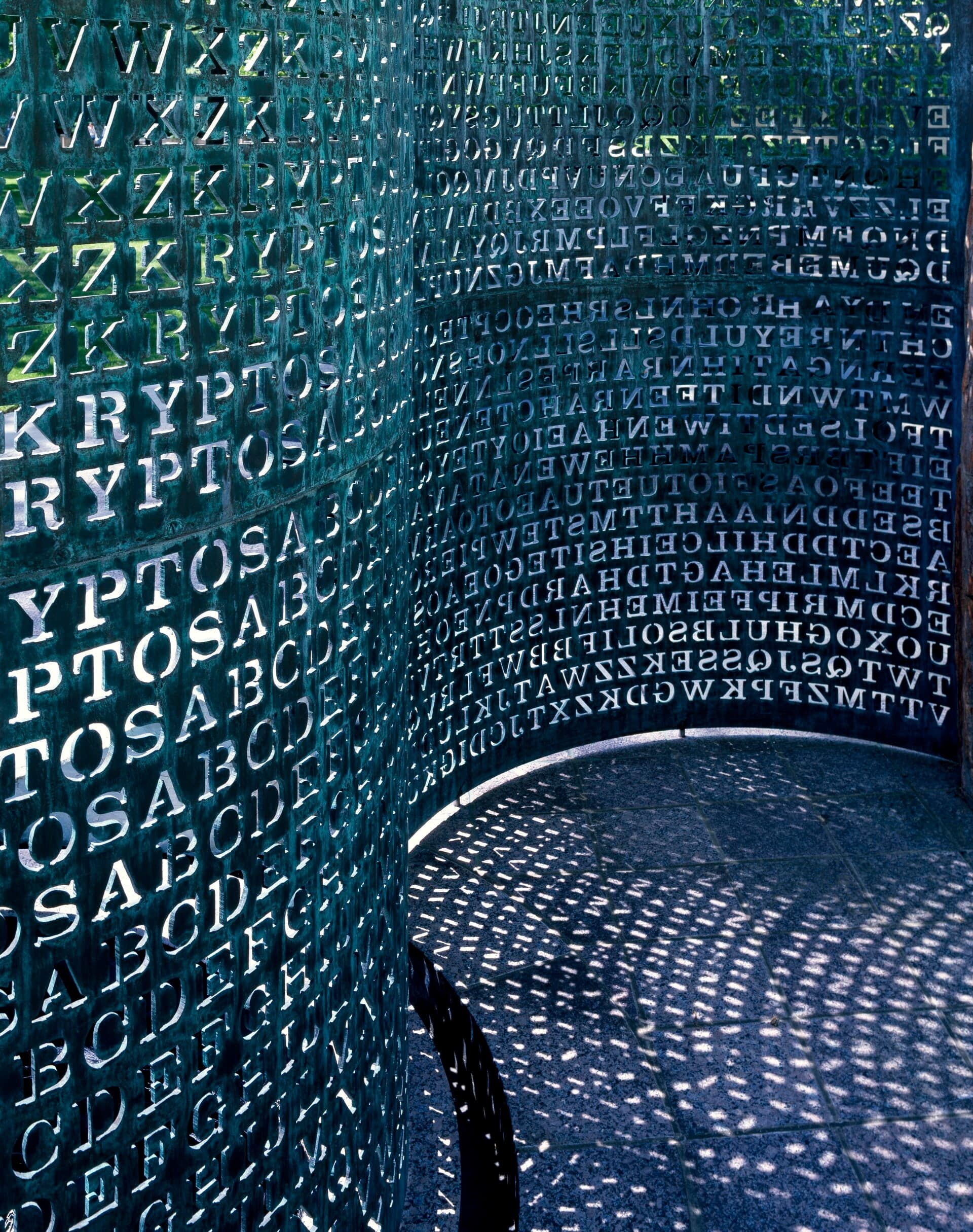 A curved copper screen perforated with rows of letters, set outdoors among dark stone, pale wood and water on a landscaped institutional site.
