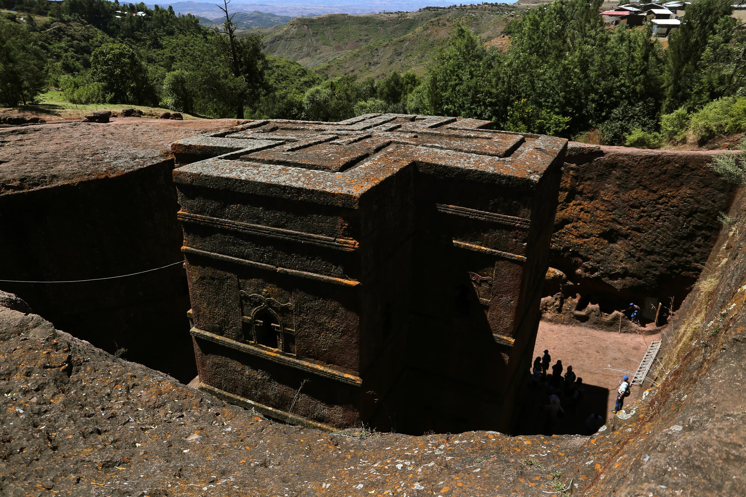 A church carved in the shape of a cross, sunk into the ground, with vertical rock walls forming a deep trench around it. The roof is flat and level with the surrounding earth.