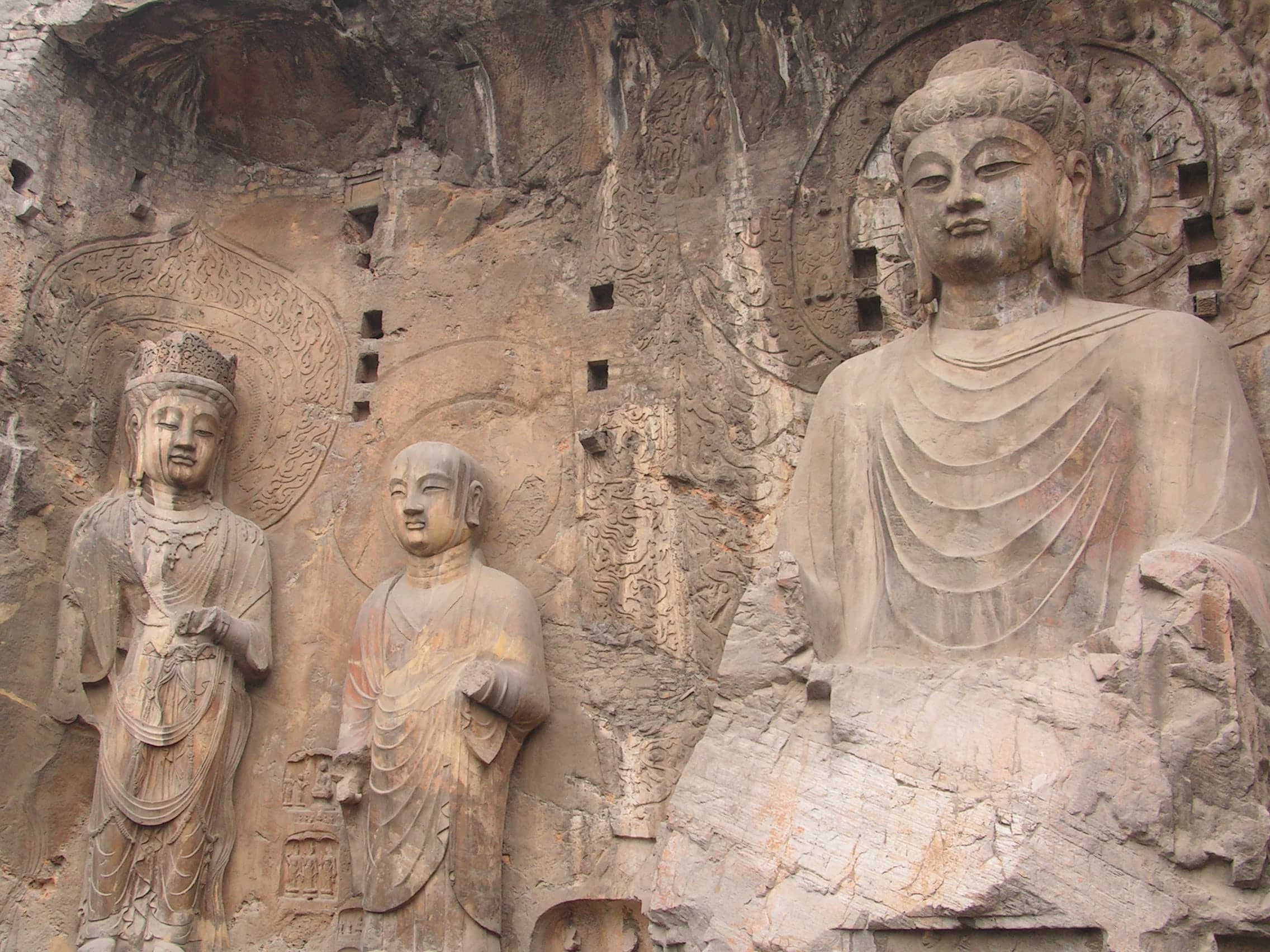A colossal seated Buddha carved into a limestone cliff, with a serene face and simplified robes. Smaller attendant figures stand to either side, dwarfed by the central figure.