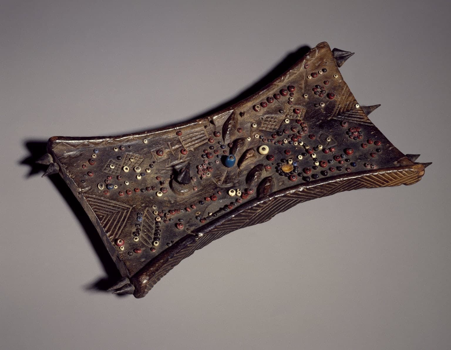 A wooden board studded with beads and metal pins in an irregular pattern, with a carved head at the top and geometric incisions visible on the surface.