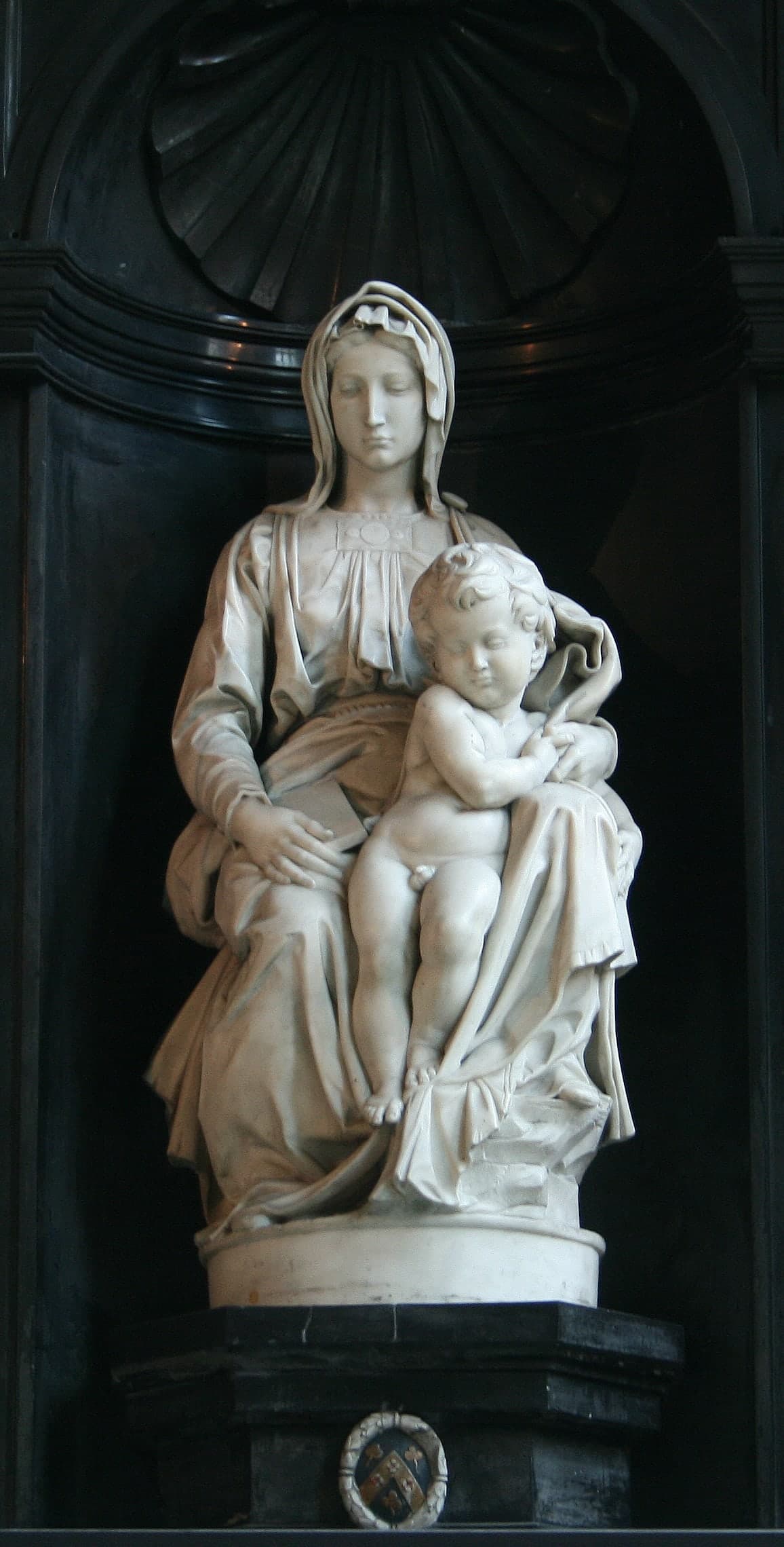 A marble Virgin seated with a standing child between her knees. She looks down and away; he faces forward, lightly held by her hand, as if about to step out from the folds of her heavy robe.