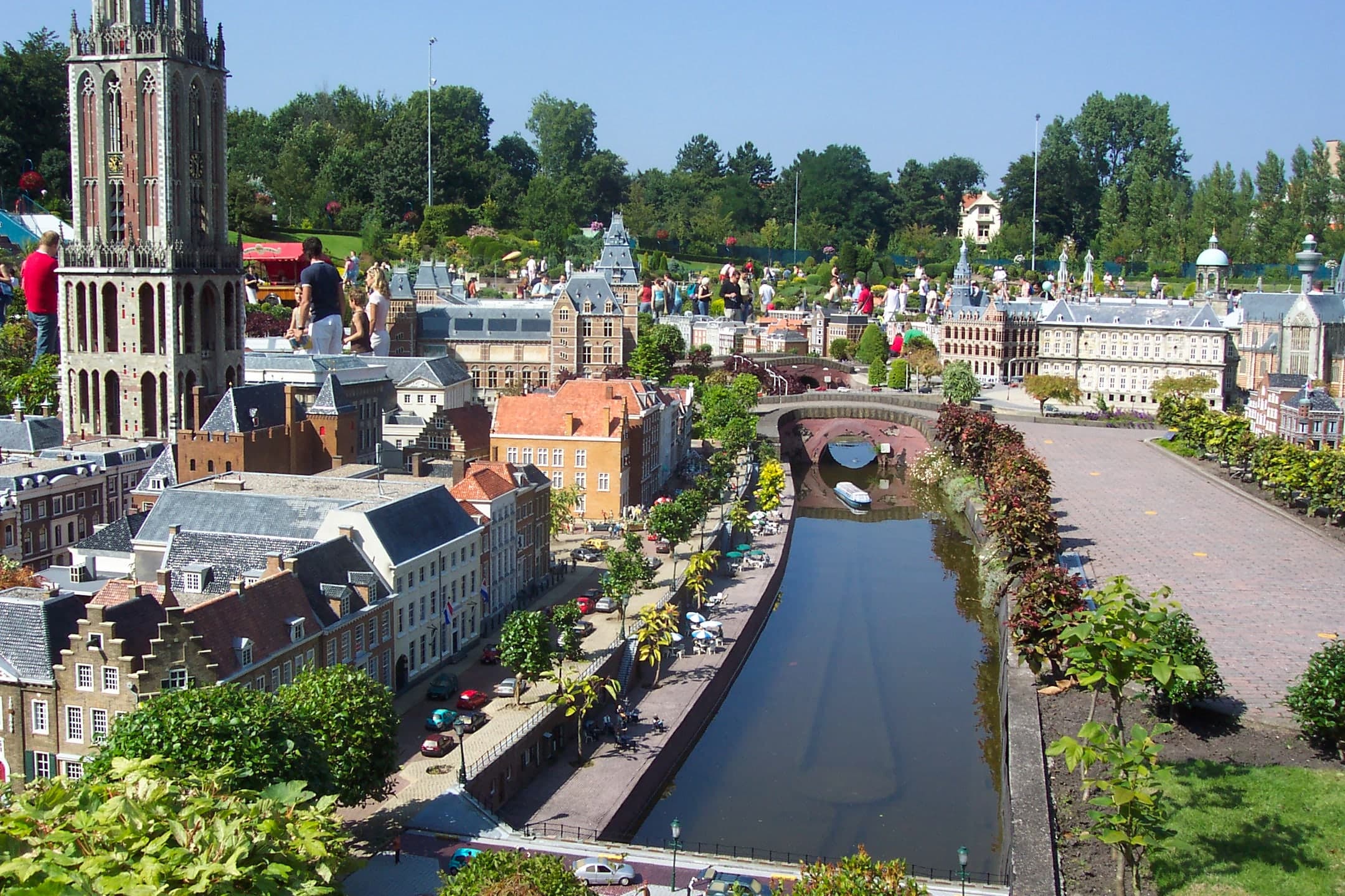 Outdoor miniature city at walking height: detailed model buildings, streets and towers under open sky, with full-size trees and paths running between the scaled-down landmarks.