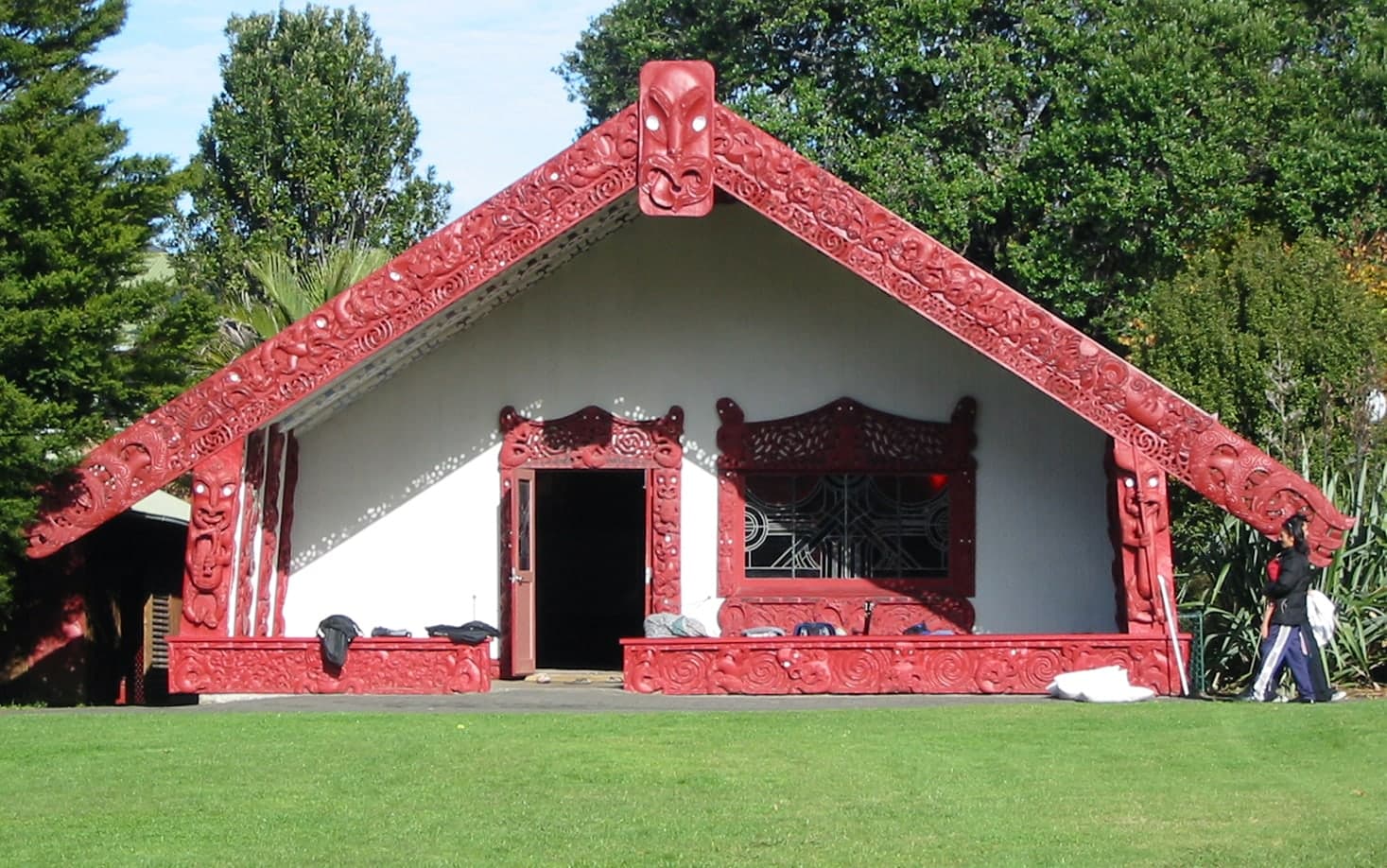 Carved wooden meeting house with steep gabled roof, ornate bargeboards extending like arms, and a carved ancestor mask at the apex. Interior walls lined with carved panels and woven geometric lattice.