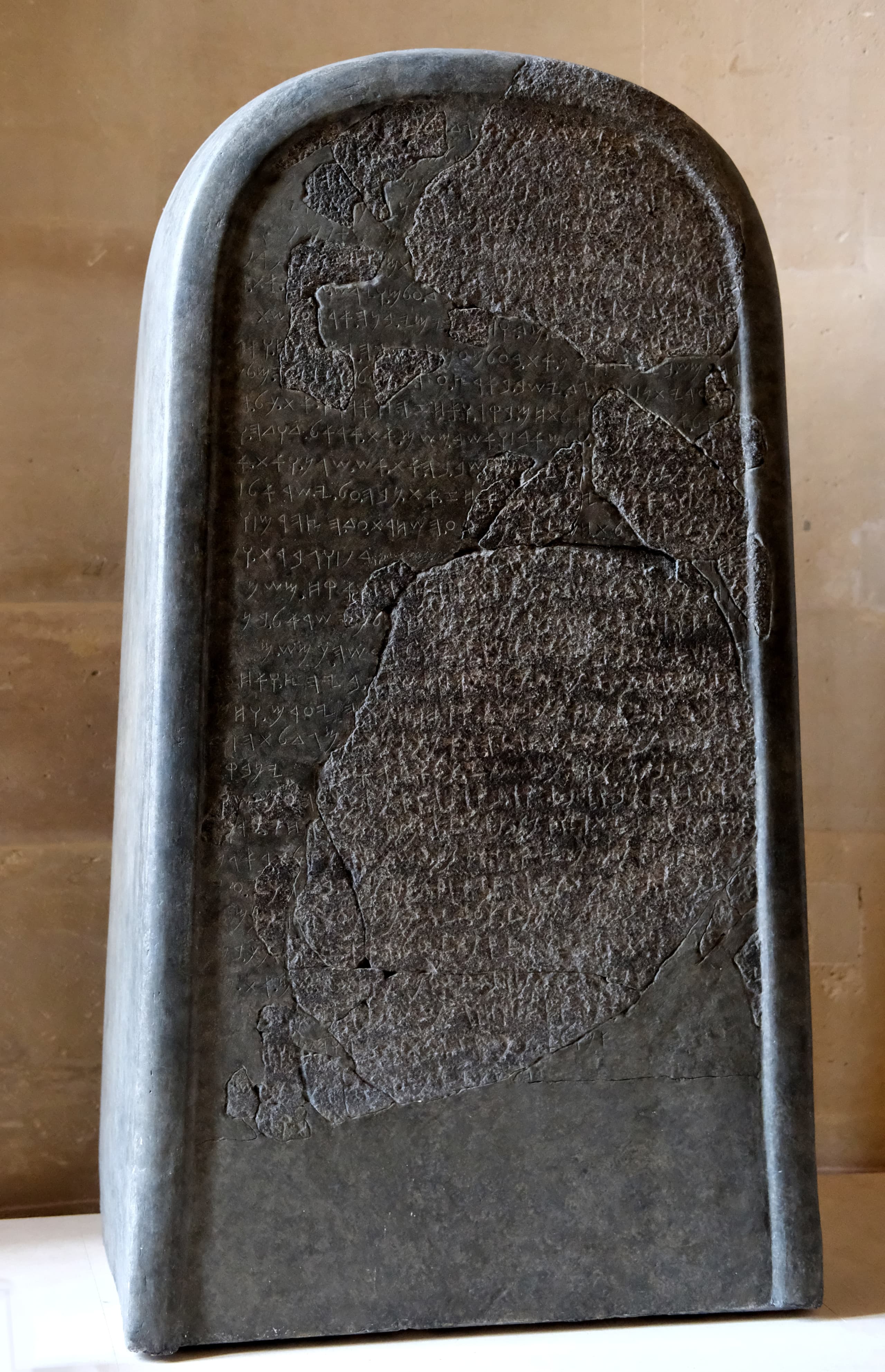 A tall, dark rectangular stone slab with horizontal lines of carved text running across its surface. The stone is broken into fragments that have been fitted back together, with visible joins and filled gaps.