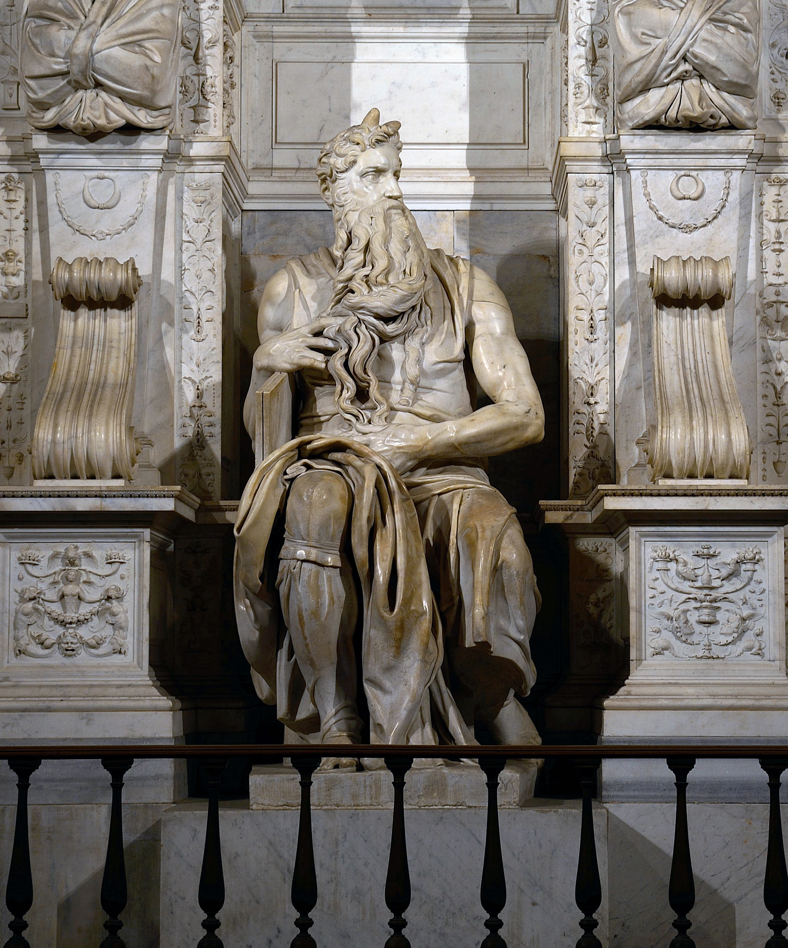 A seated bearded figure carved in marble, head turned sharply to the left, with two short horns rising from the forehead. One hand rests across the chest, fingers in the beard.