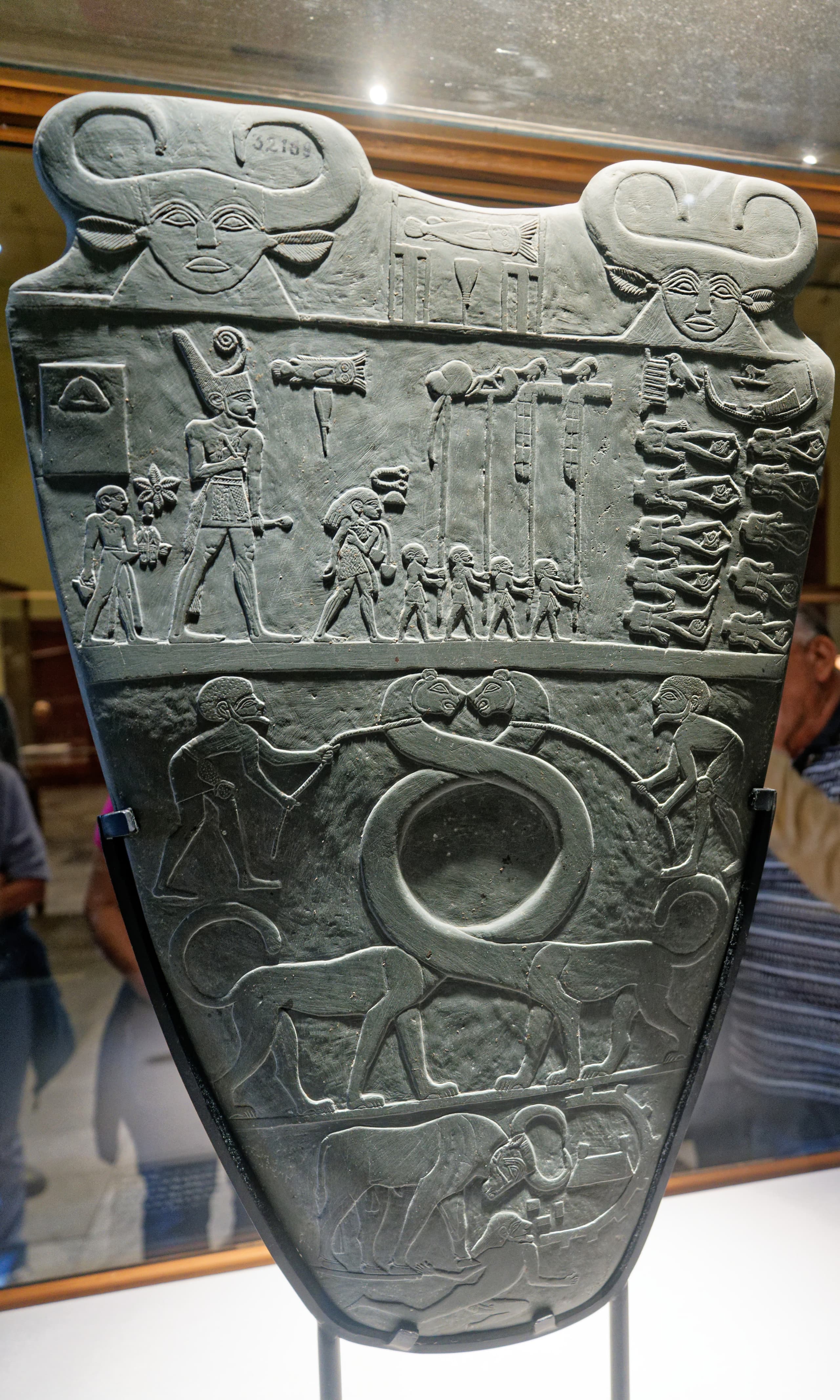 A tall, shield-shaped slab of dark stone carved on both sides with figures in low relief. A large central figure in a kilt and crown dominates each side, surrounded by smaller human and animal figures arranged in horizontal bands.