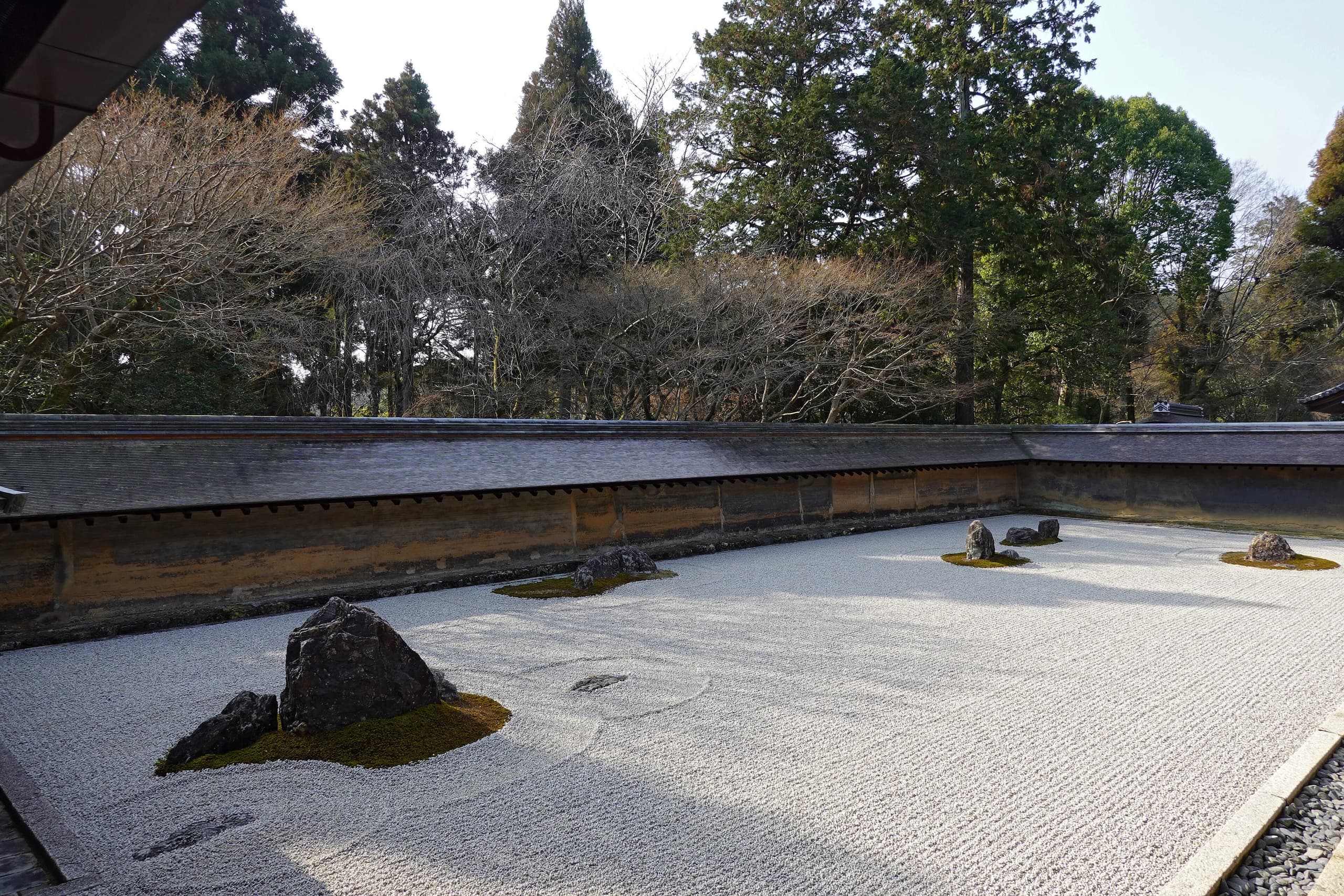 A rectangular enclosure of white raked gravel containing fifteen dark stones arranged in five asymmetrical groups, bounded by a low weathered earthen wall.