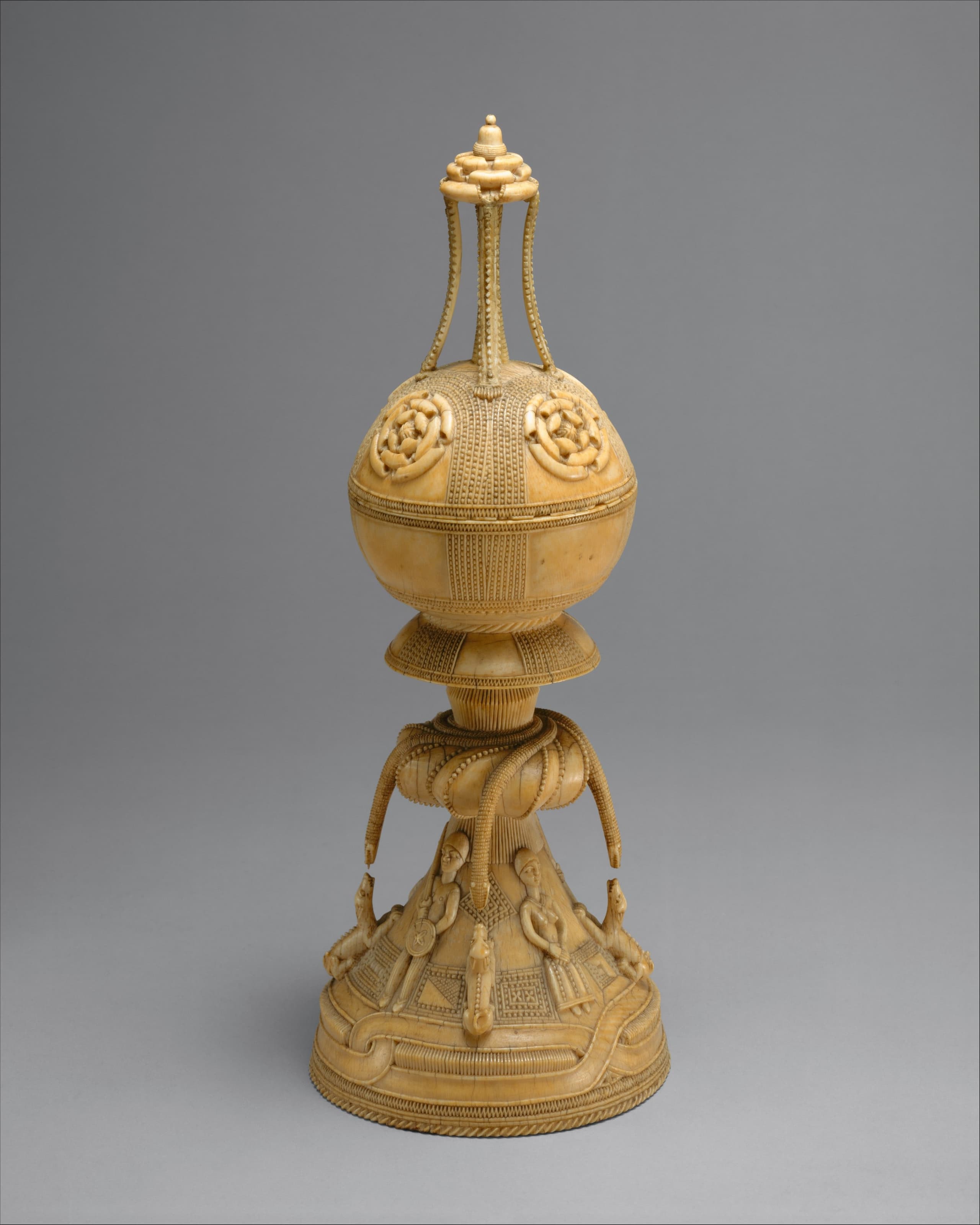 A carved ivory vessel in two tiers, with a lid. Figures in European dress stand around the lower section. Geometric patterns band the surface. A small carved shape tops the lid.