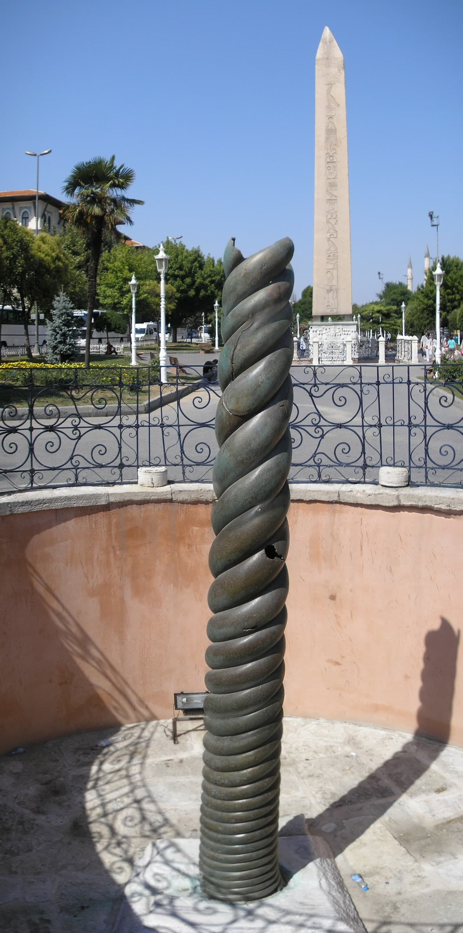 A dark, weathered bronze shaft standing outdoors, its surface formed by three serpents coiled in a continuous spiral, truncated at the top where the heads are missing.