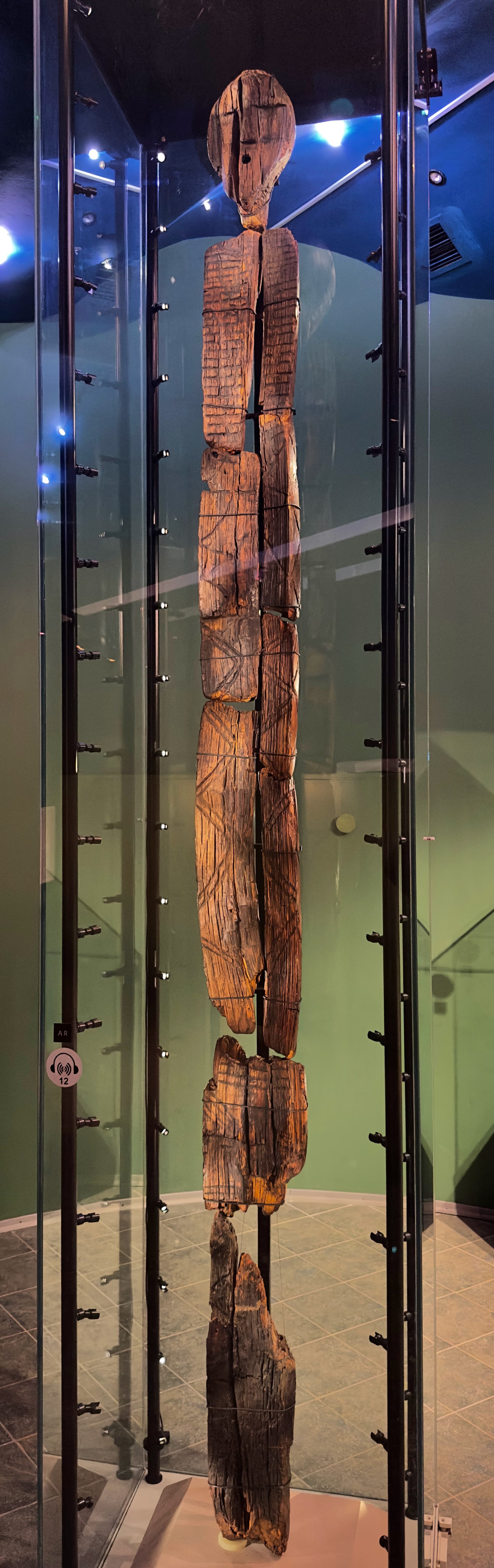 A tall, dark wooden figure with a carved human head at the top and a long shaft marked by horizontal bands, angles and further face-like features.