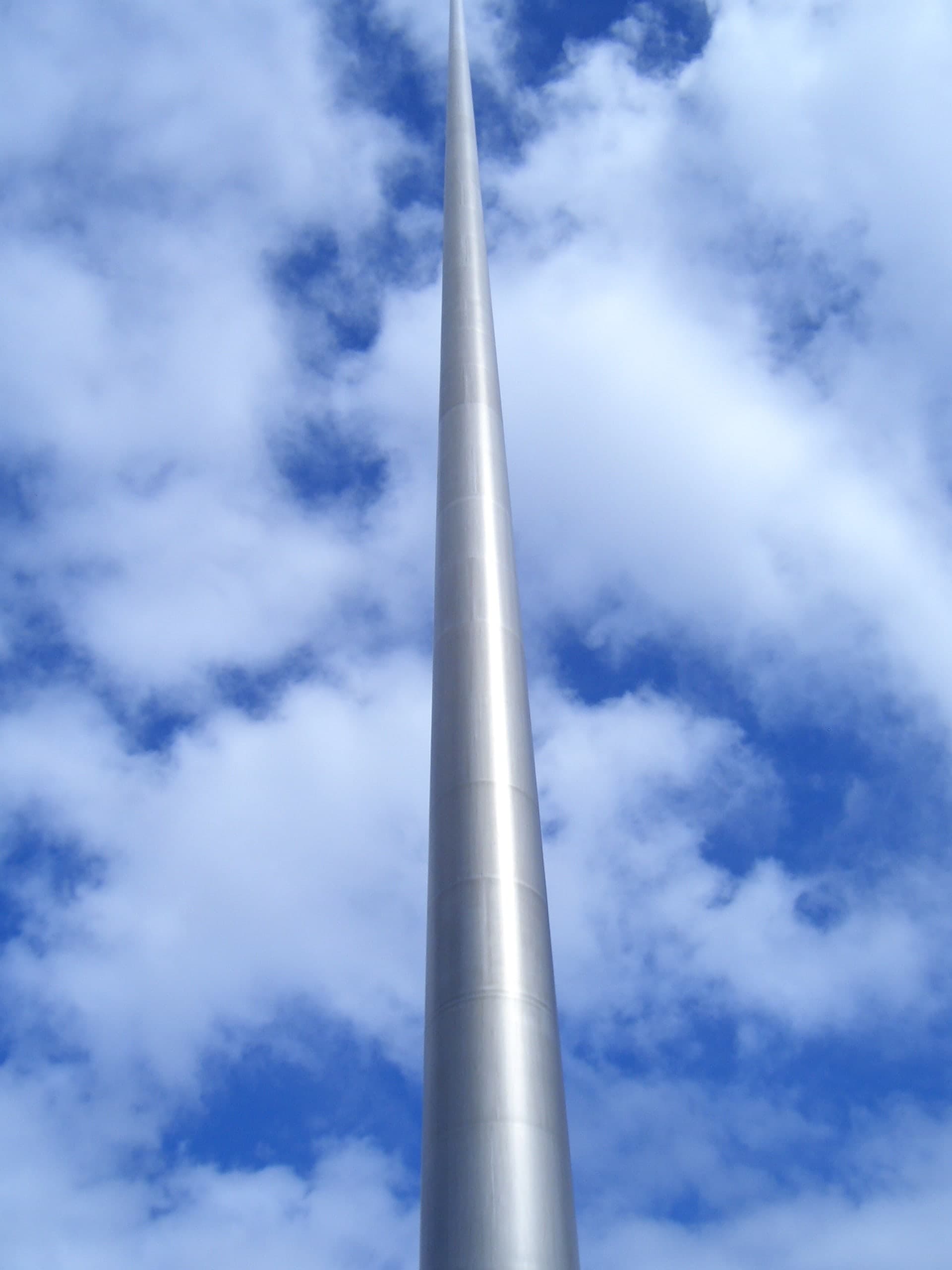 A tall, slender pin-like shaft of pale steel rising above a wide city street, tapering to a fine point against the sky, with surrounding buildings much lower than its tip.