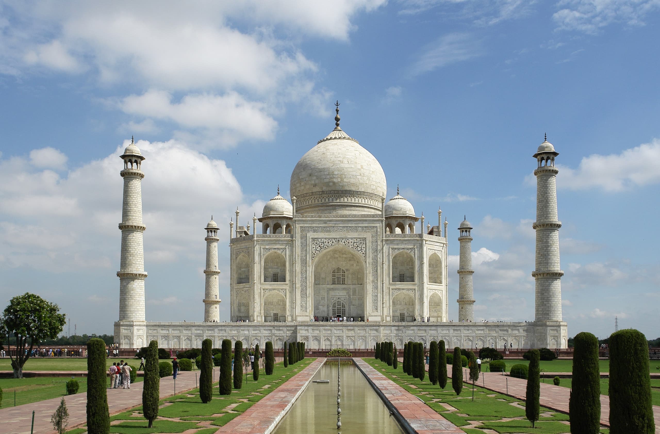 A white marble mausoleum with a central dome and four minarets, reflected in a long rectangular pool lined with dark trees.