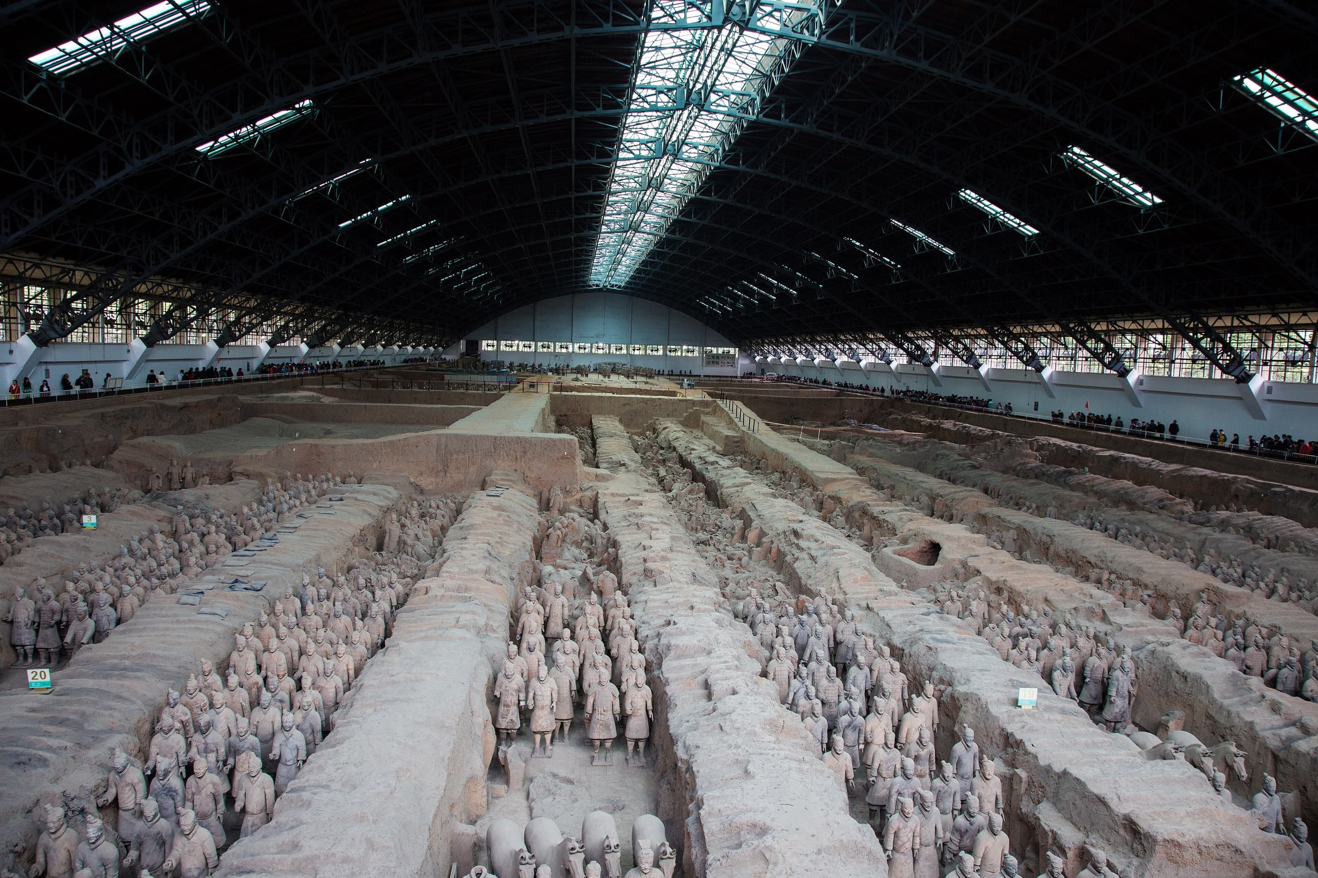 Rows of life-sized terracotta figures standing in excavated earth pits, each with distinct facial features, dressed in armoured tunics, their surfaces a uniform grey.
