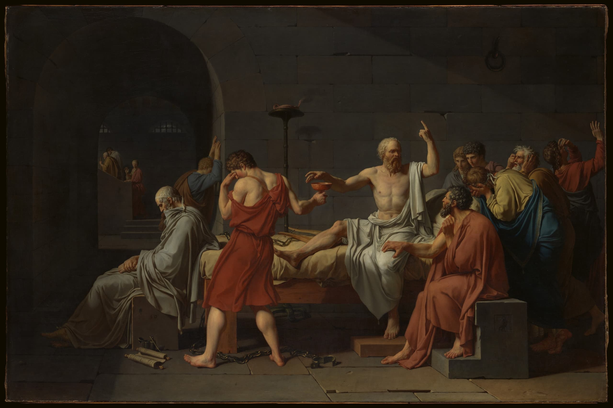 A bare prison room. A muscular older man sits upright on a bed, one hand raised in argument, the other reaching for a small cup held out by a turning figure. Around him companions gesture, lean in, or cover their faces. Open chains lie on the stone floor.