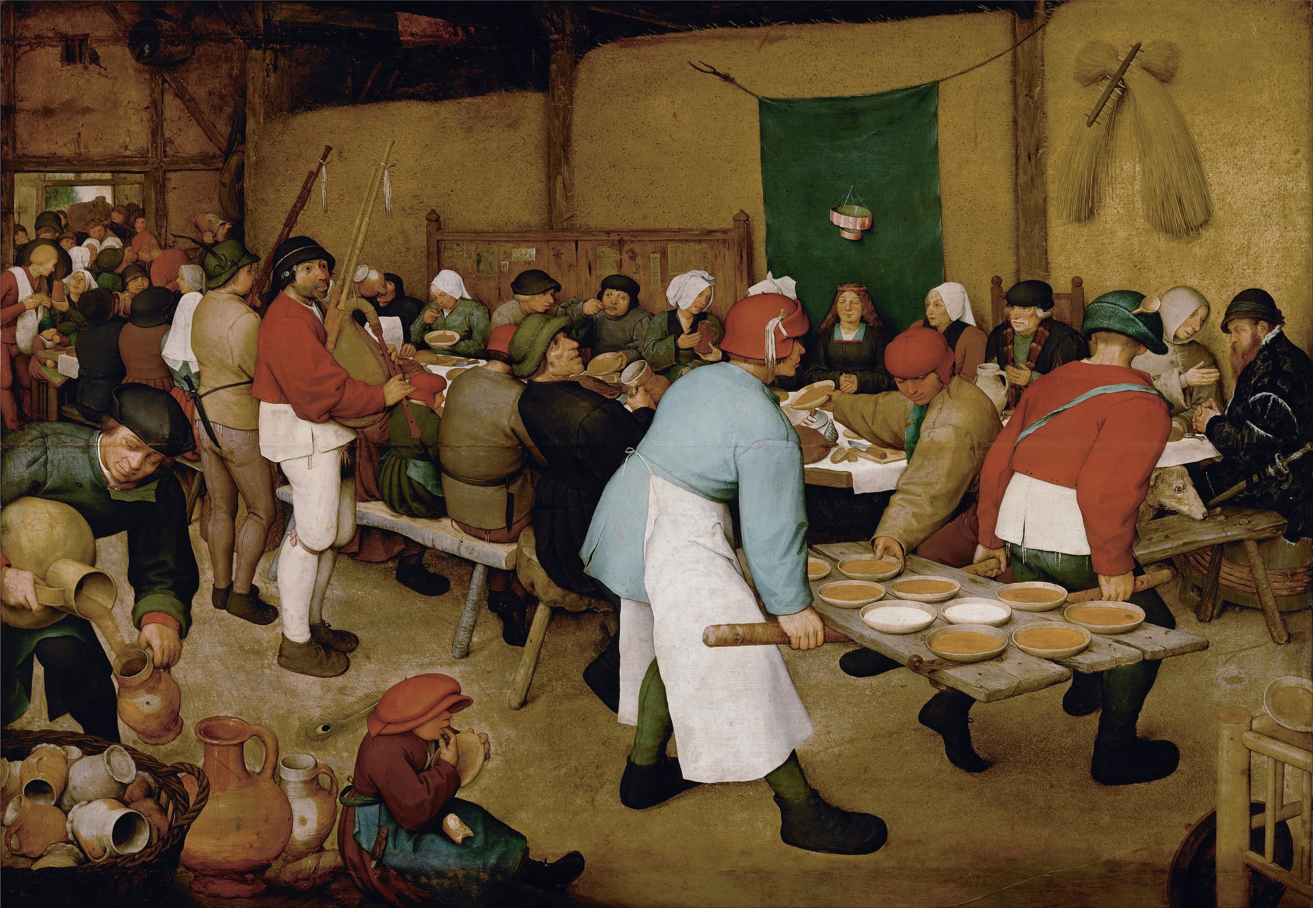 A crowded barn interior. Guests eat at a table running diagonally into shadow; two men carry a wide board of bowls forward. A young woman sits still against a dark hanging under a small paper crown, with stacked sheaves of grain behind.