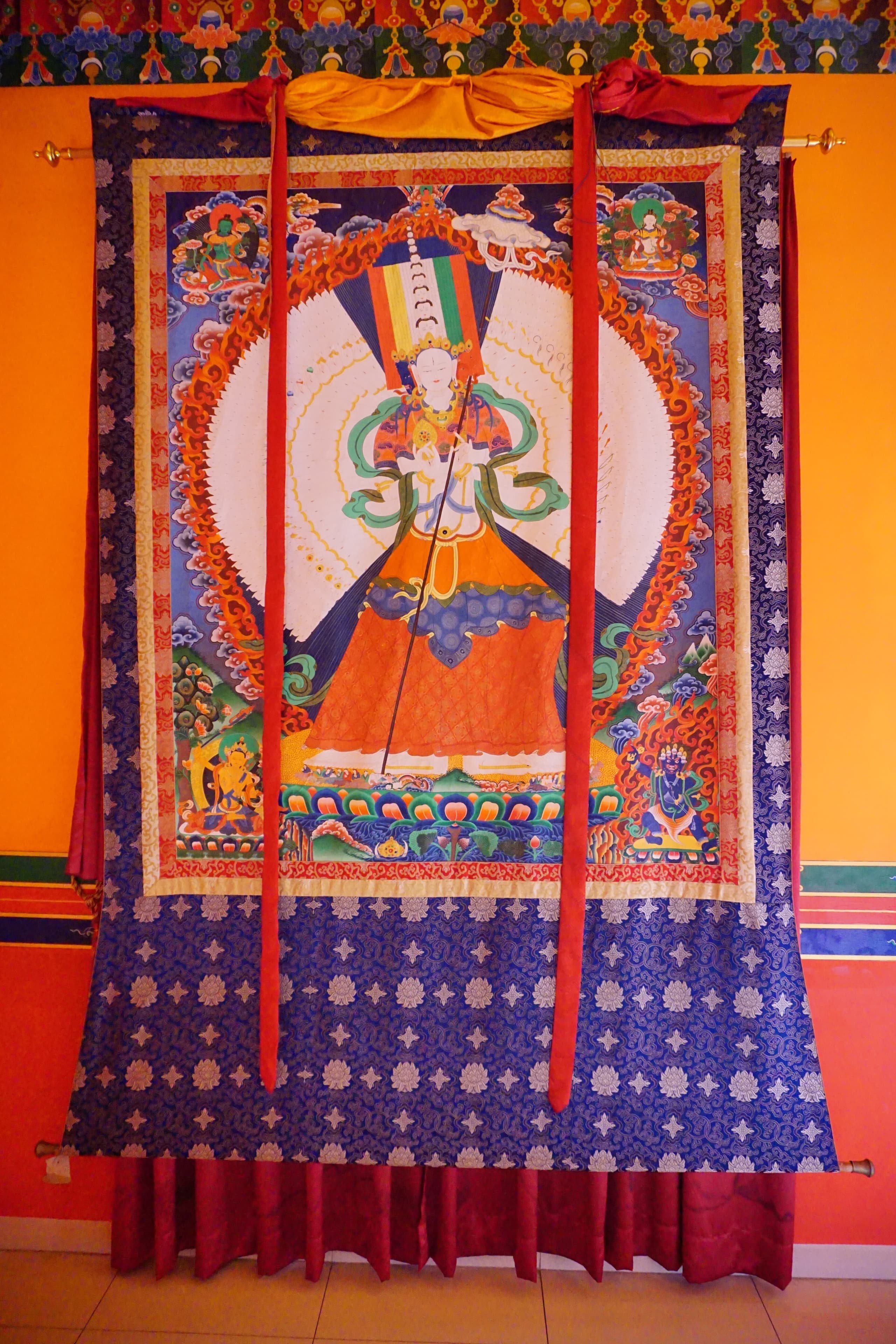 A Tibetan Buddhist painting on cloth showing a central deity surrounded by smaller figures arranged symmetrically, with rich mineral colours and gold detailing.