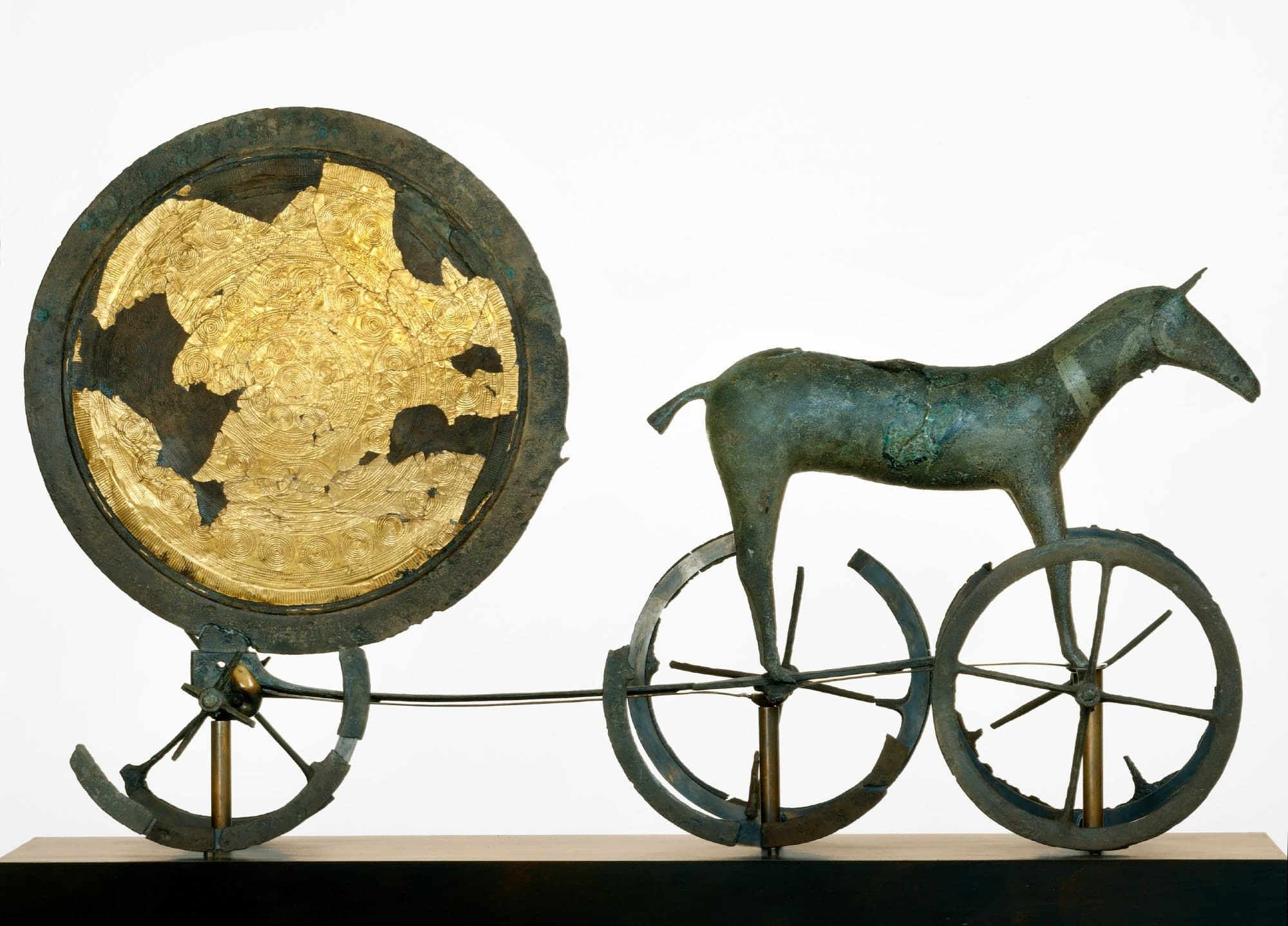 A small bronze horse stands before a large round disk finished with gold, the pair mounted on a low carriage with spoked wheels.