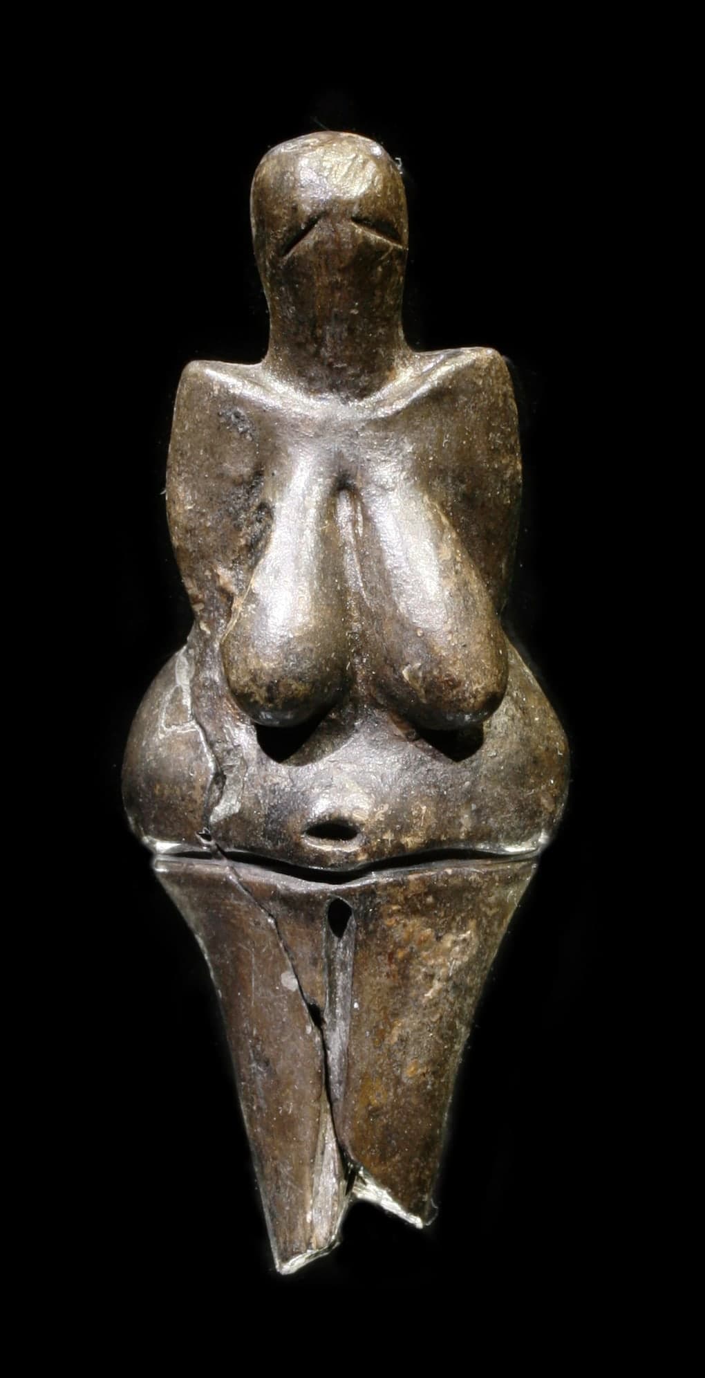 A small brownish ceramic statuette of a nude woman, hips and breasts enlarged, head reduced, arms close to the body, legs short and tapering, shown against a plain background.