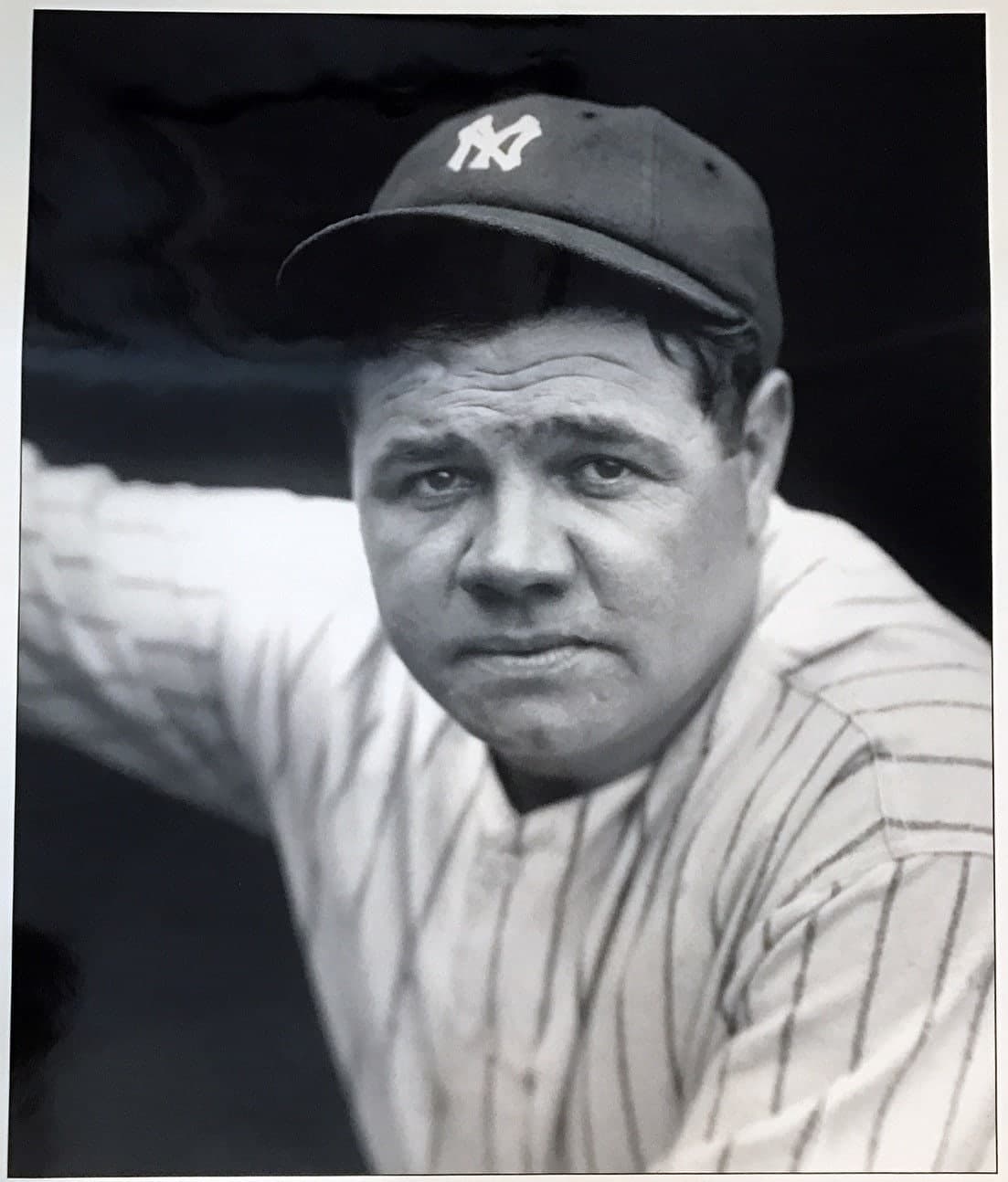 Black-and-white photograph of a man in a light-coloured baseball uniform with dark pinstripes, a cap, and a serious expression, holding a bat upright beside him.