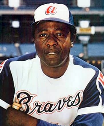 Head-and-shoulders portrait of a smiling man in a white baseball uniform with navy sleeves and red piping, a white cap with a red letter on it, a bat resting against his shoulder and empty stadium seating behind him.