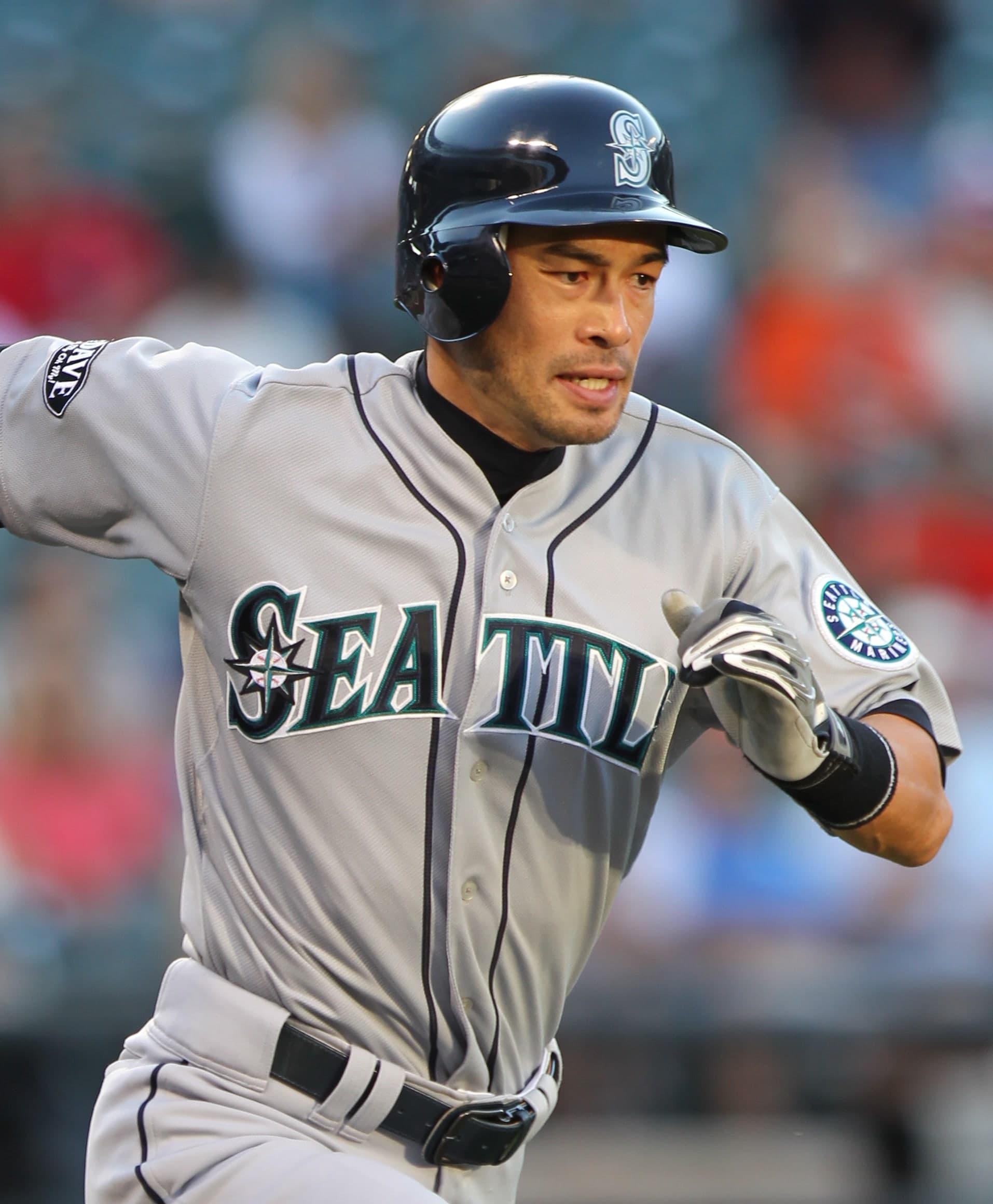 A baseball player in a dark blue cap and grey uniform with blue and teal trim, holding a bat in a ready stance, looking toward the pitcher.