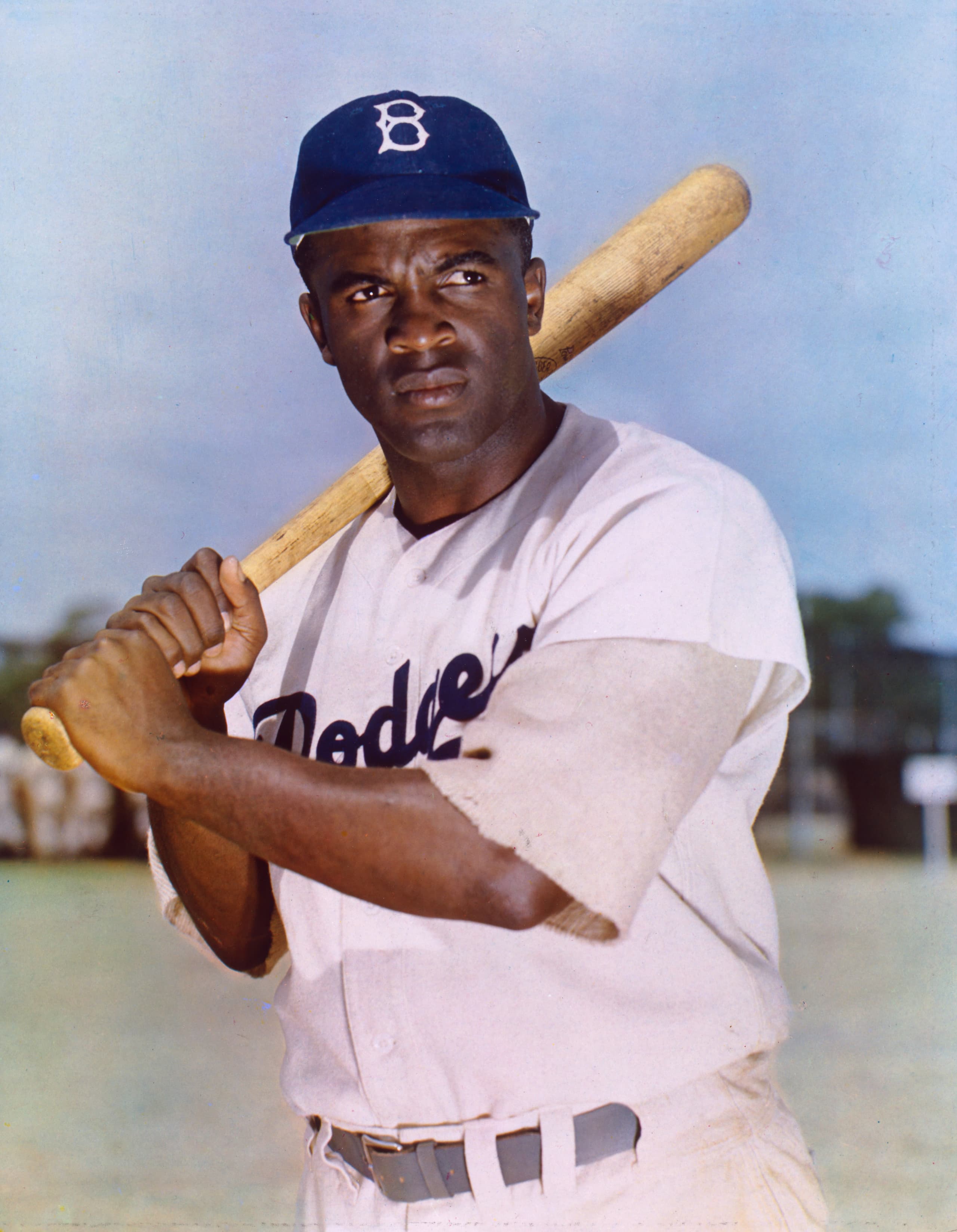 A baseball player in a white uniform with 'Dodgers' across the chest, wearing a cap with a 'B' on it, crouched in a fielding stance on a dirt infield with empty stadium seats visible behind him.