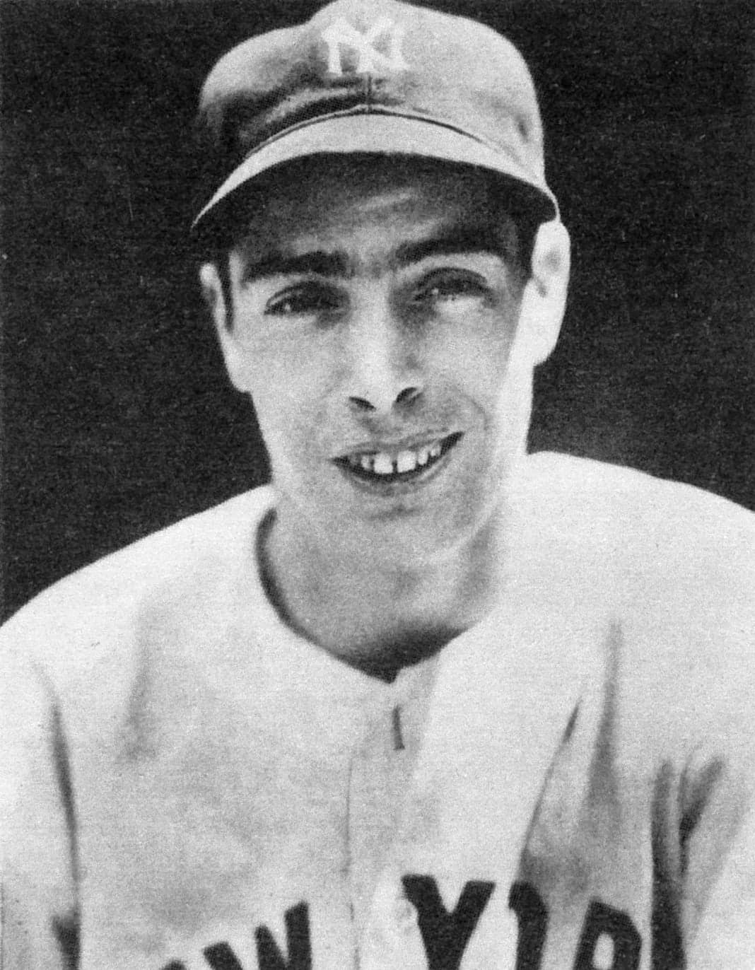 Black-and-white photograph of a man in a pinstriped baseball uniform and cap, holding a bat across his shoulders with both hands, standing on a grass field.