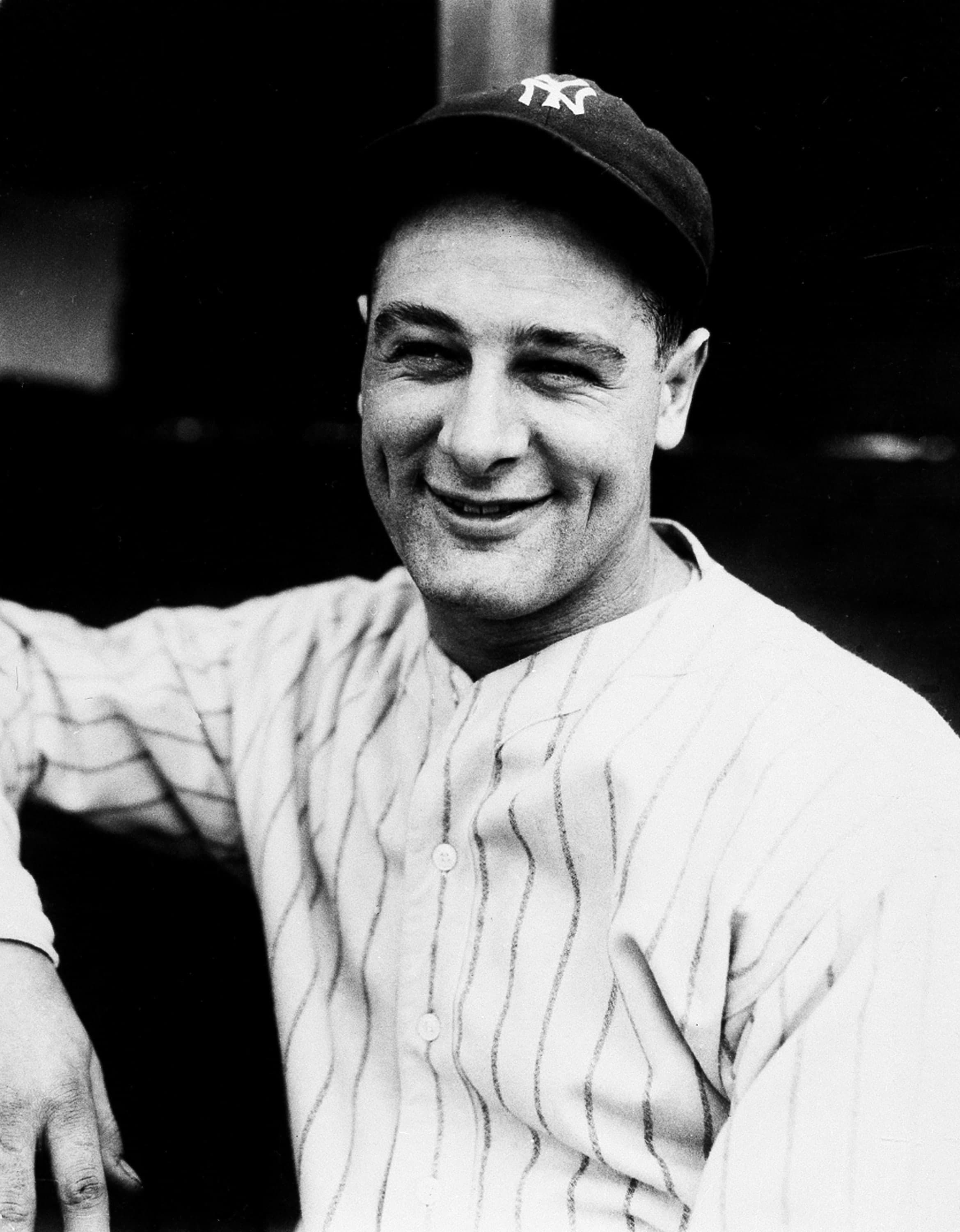 Black-and-white photograph of a baseball player in a pinstriped uniform and cap, holding a bat across his shoulders with both hands, standing on a field with empty stadium seating visible behind him.