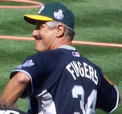 A baseball pitcher in mid-delivery on the mound, wearing a white uniform with a distinctive handlebar moustache, right arm extended forward after releasing the ball.