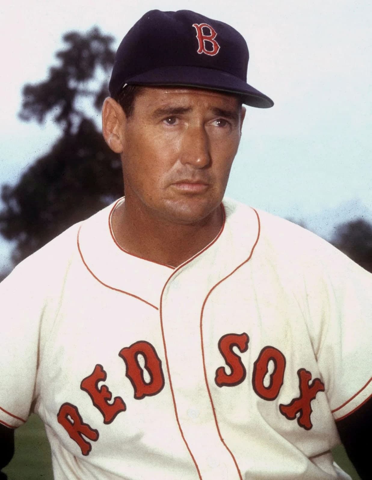 A baseball player in a dark cap and light uniform stands holding a bat, his weight on one leg, looking slightly to the side with a calm expression.