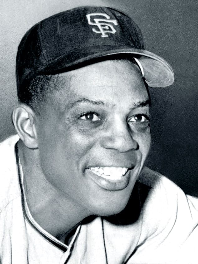 Head-and-shoulders portrait of a smiling man in a white baseball uniform with dark sleeves, a white cap with a dark letter, a bat resting against his shoulder and empty stadium seating behind him.